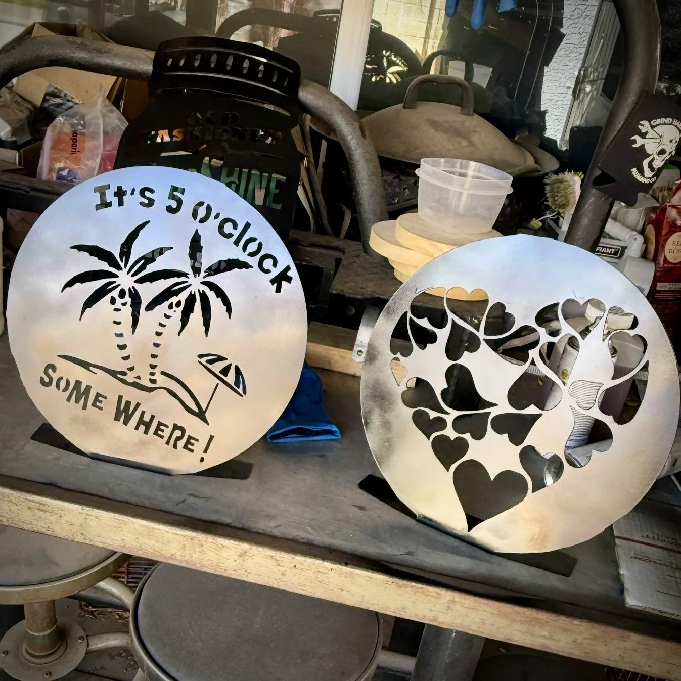 Two round metal signs with decorative cutouts standing on a table inside a workshop. The left sign features two palm trees, a beach umbrella, and the text 'It's 5 o'clock somewhere!' The right sign has a cutout of a drink with a straw, surrounded by 