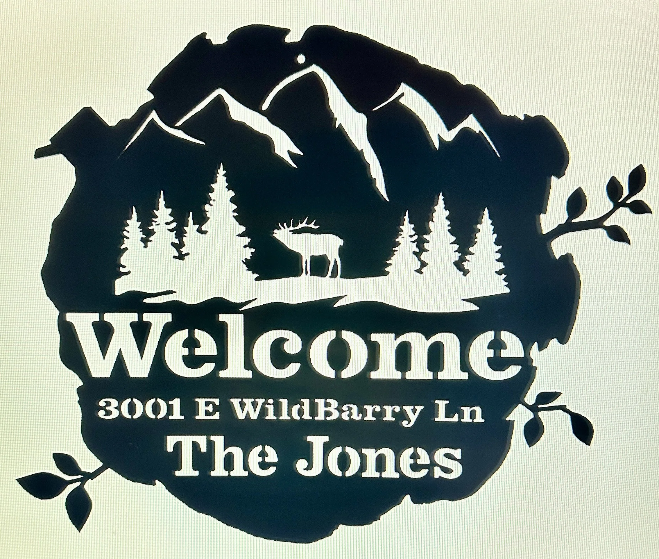 A black cutout sign features a mountain range, pine trees, a bear, and a branch with leaves above the text. The text reads 'Welcome' and below it provides an address: '3001 E WildBurry Ln'. The sign also states 'The Jones'.