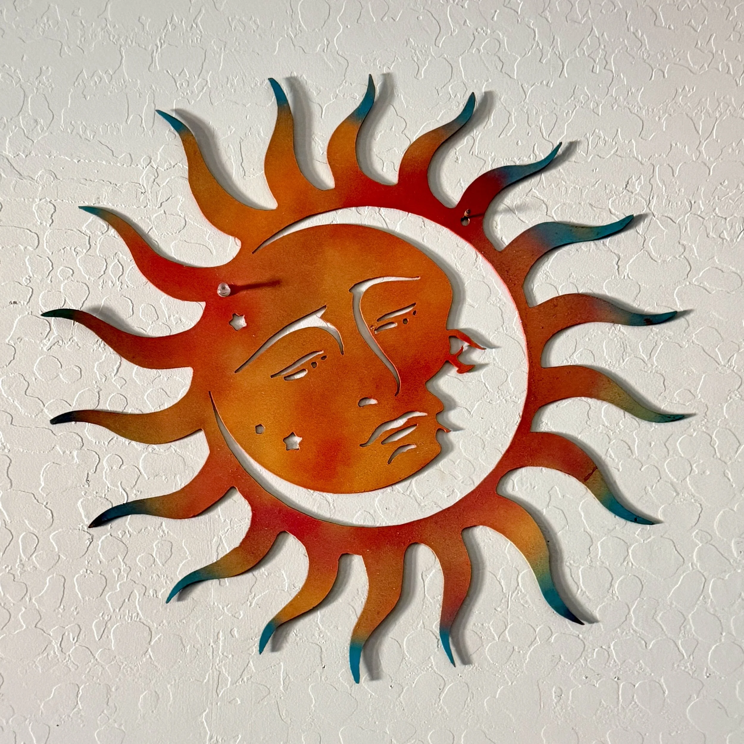 Colorful metal sun decoration with rays extending outward, mounted on a textured white wall.