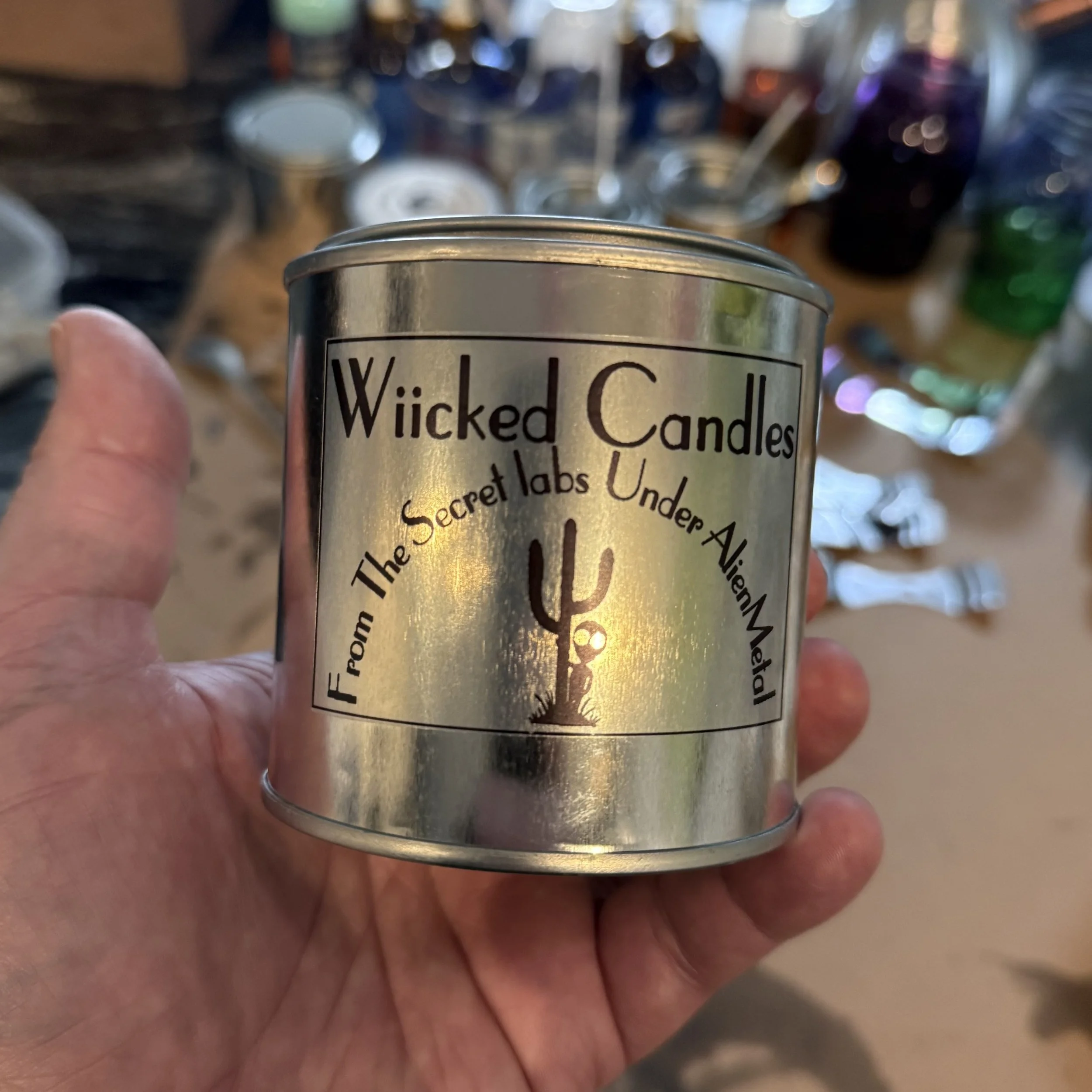 Close-up of a hand holding a metal can labeled 'Wicked Candles' with a blurred background of various bottles and objects.
