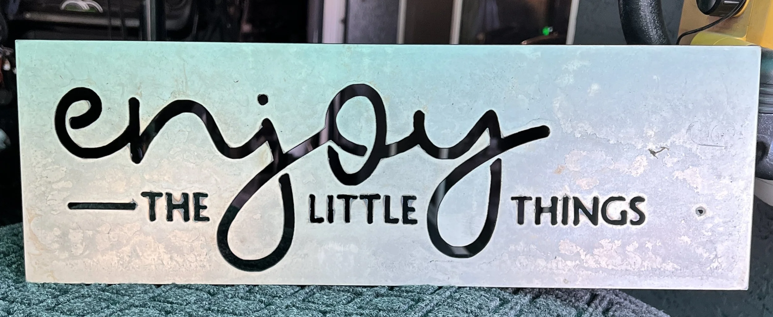 Decorative sign with cut-out words that say 'enjoy the little things' in black cursive and block lettering, set against a light-colored background.