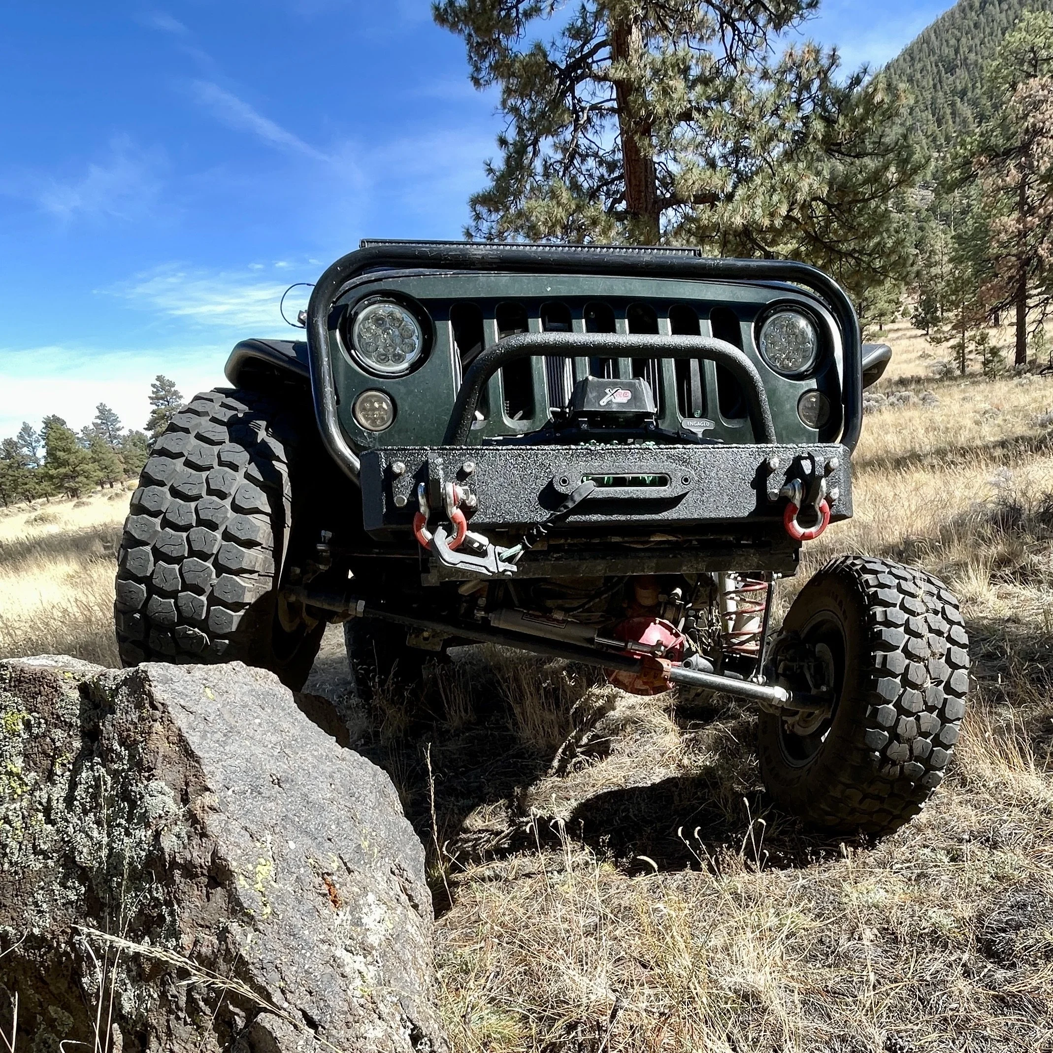 Close-up of the front of a dark green Jeep Wrangler with large off-road tires, set in a mountainous forest area with a blue sky, trees, and rocky terrain in the background.