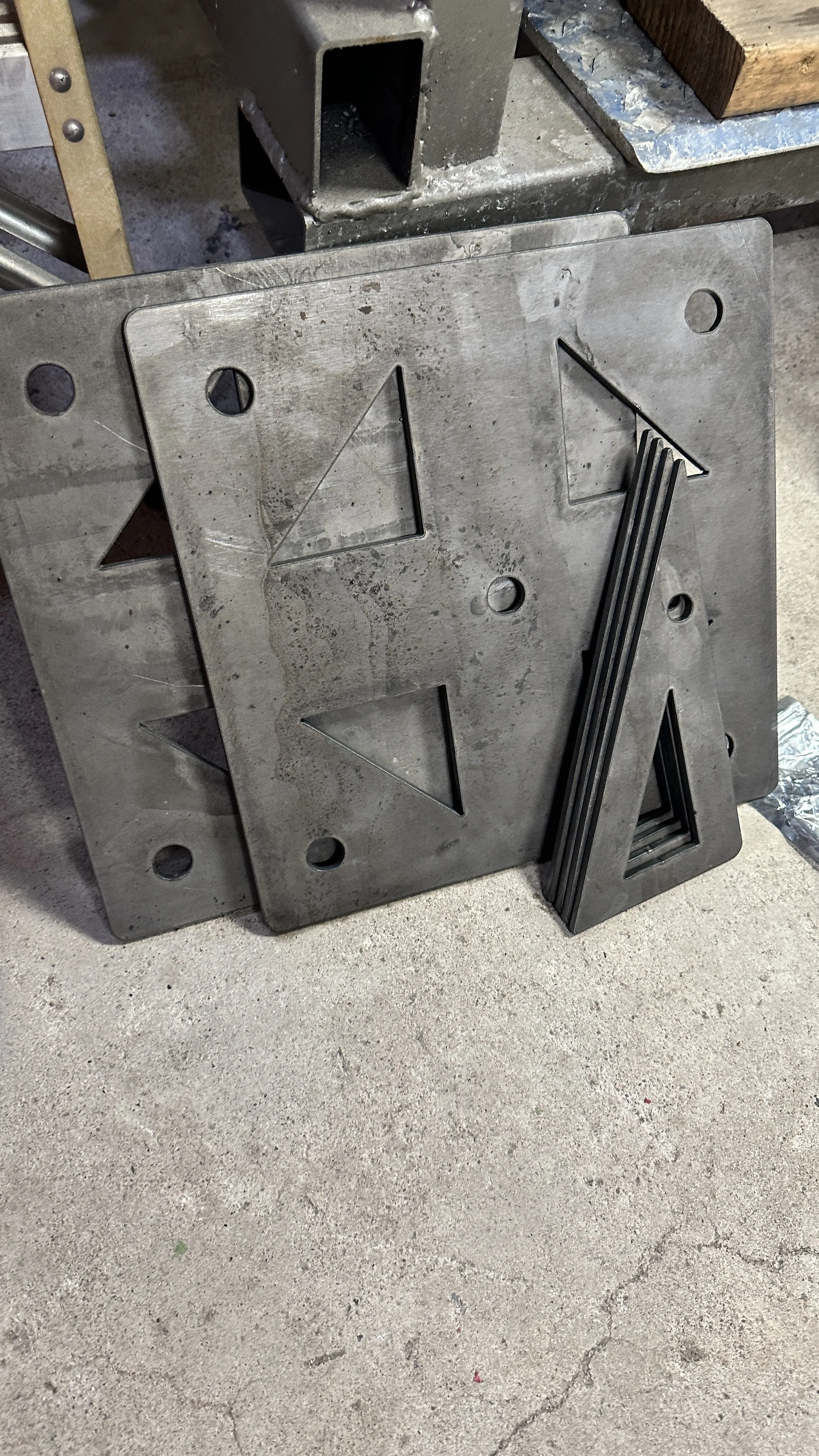 Metal cutting templates with triangular cutouts and a metal square ruler, resting against a concrete floor near a metal workbench in a workshop.