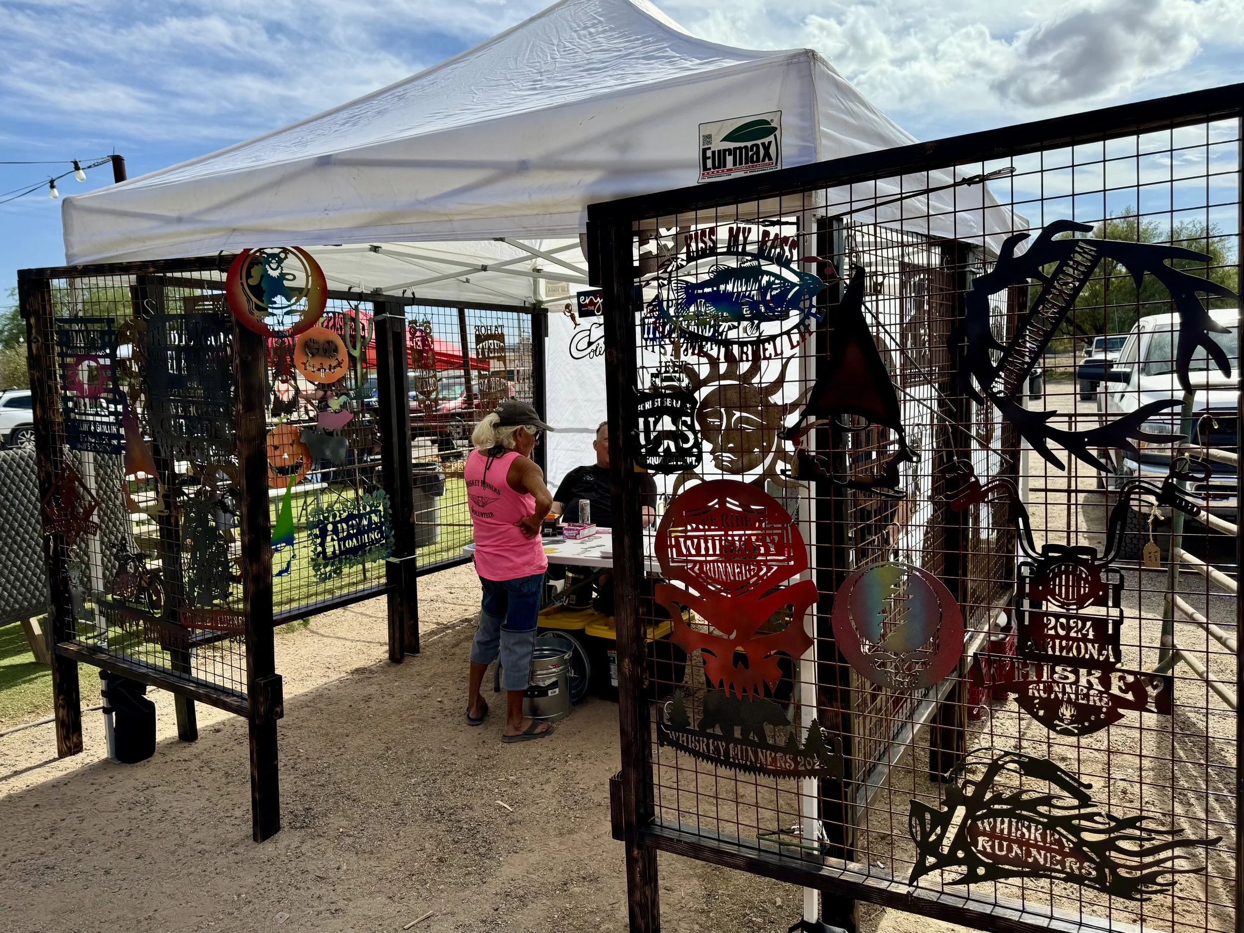 An outdoor craft fair booth with metal cutouts and signs for whiskey runners, a white canopy tent, and people browsing the items on display, with vehicles parked in the background under a partly cloudy sky.