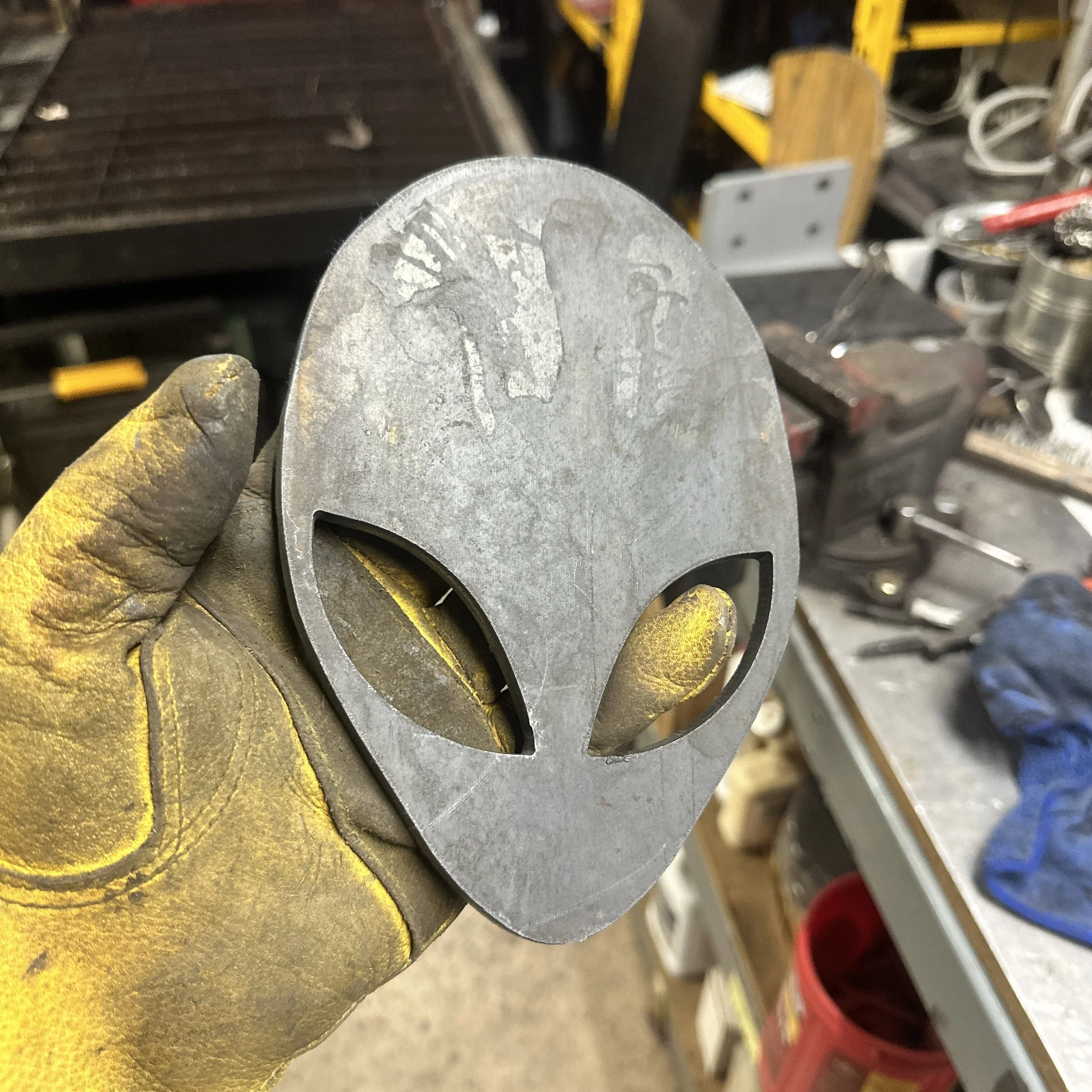 Close-up of a metallic object with engraved markings or text, held by a person wearing a yellow protective glove, in a workshop or industrial setting with tools and equipment in the background.