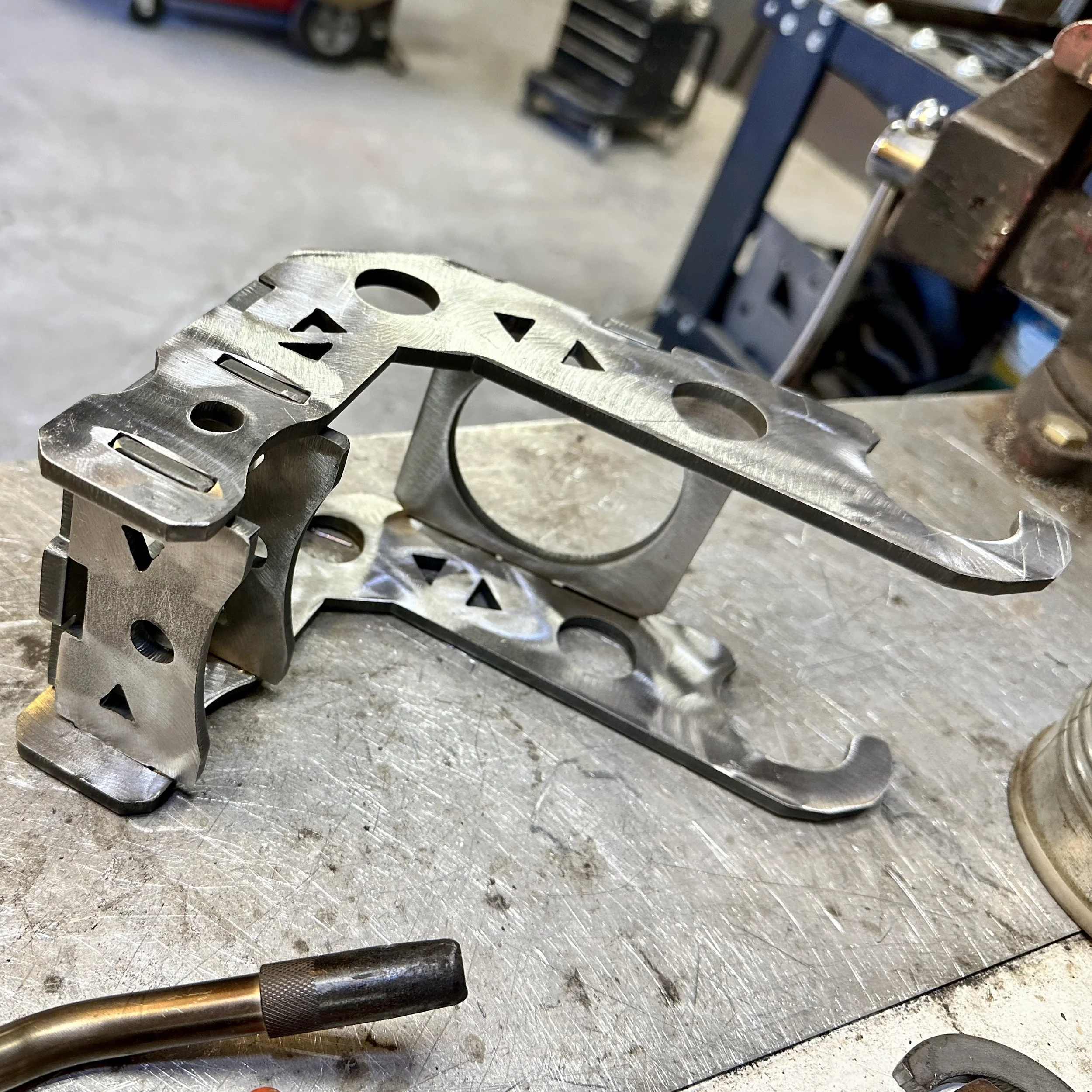 Close-up of a metal component with cutouts and a counterbore, placed on a workbench, with a welding or soldering tool nearby, in a workshop setting.
