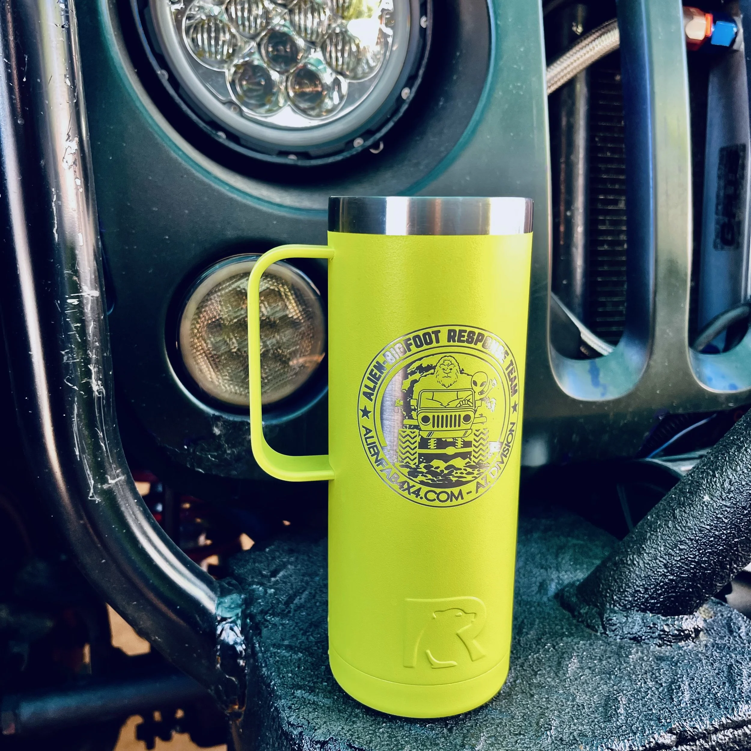 Close-up photo of a yellow travel mug with a logo, placed in front of a large LED headlight on a vehicle, with a textured green surface beneath.