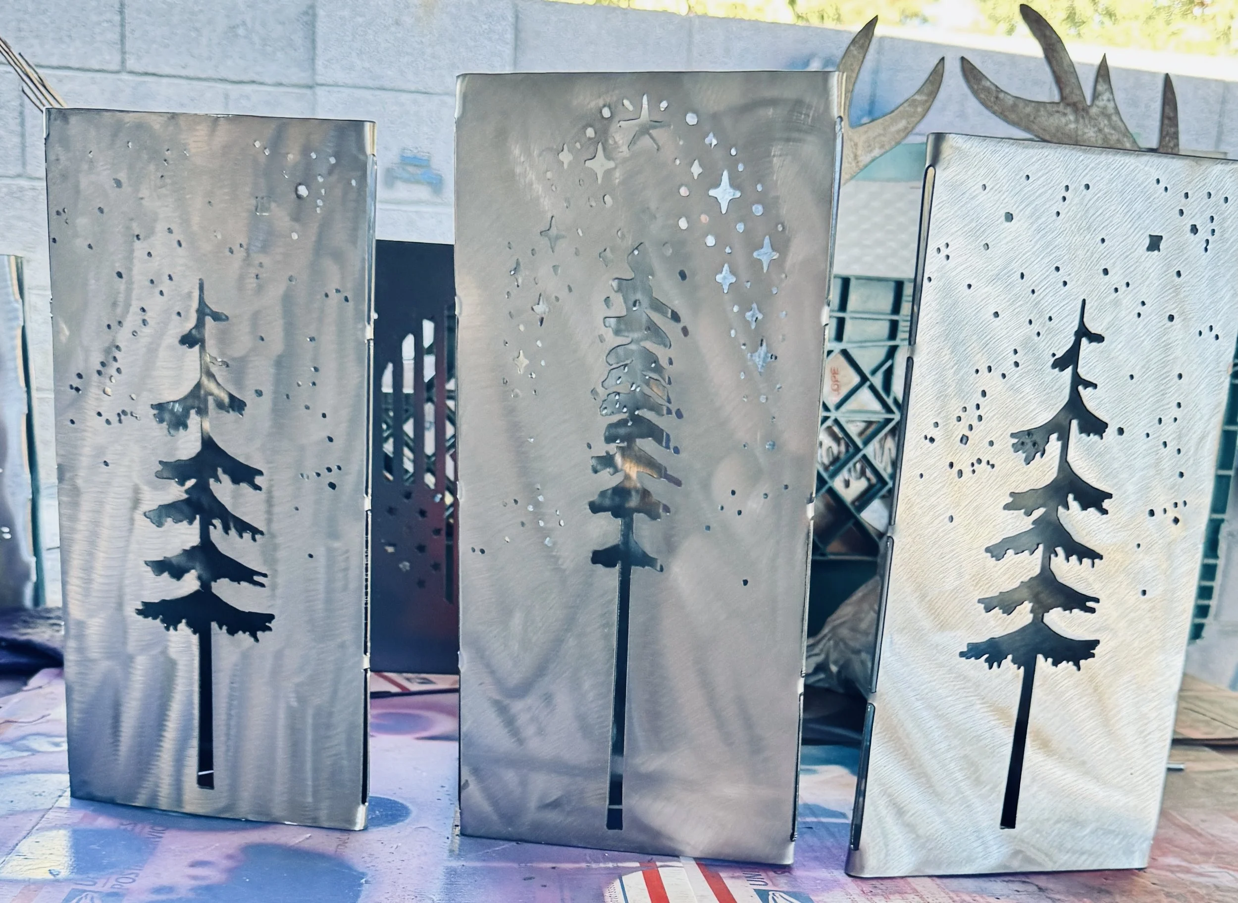 Set of four metal lanterns with Christmas tree cutout designs, arranged outdoors on a surface with a brick wall and trees in the background.