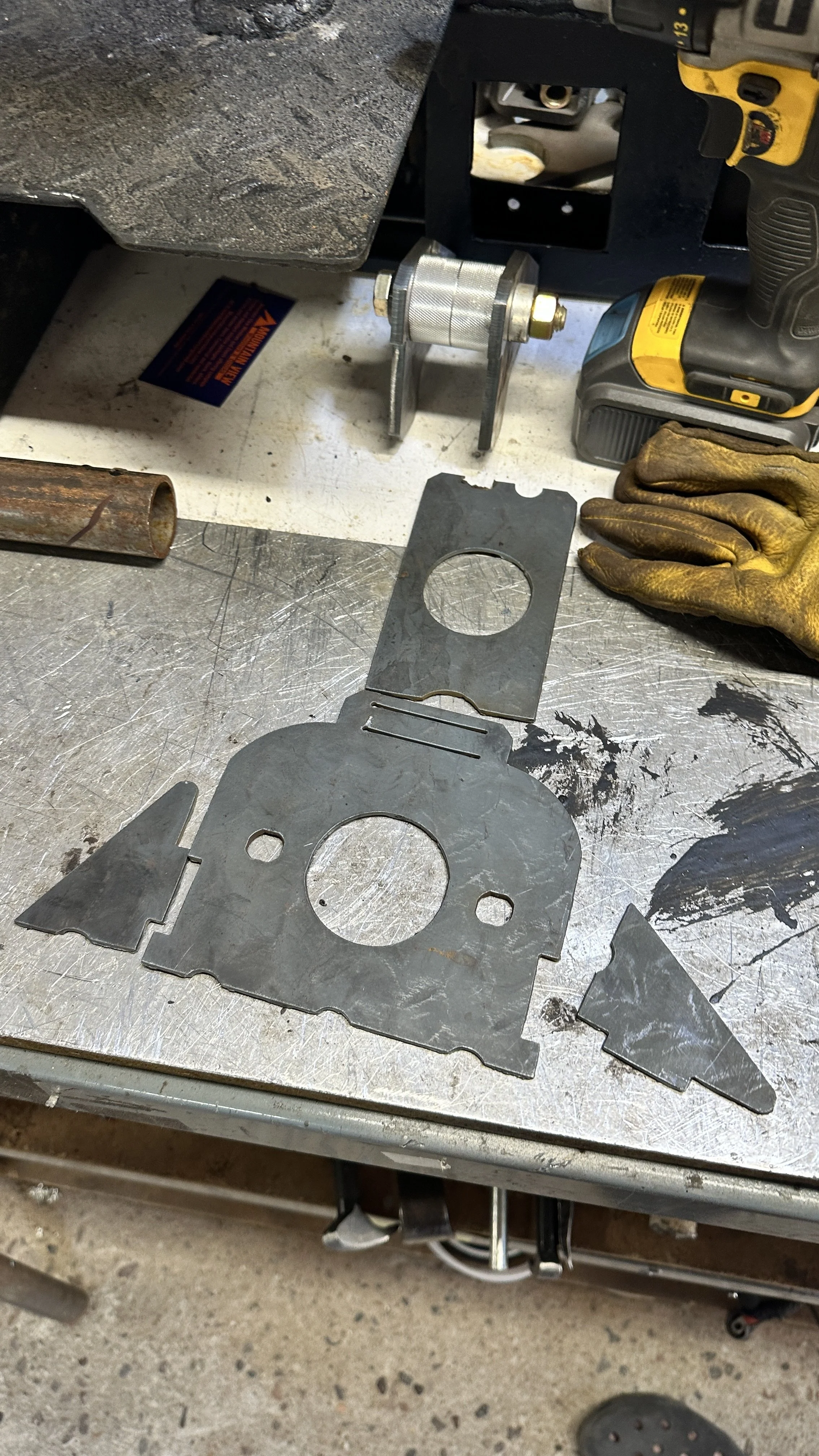 Metallic brackets and metal sheet on a workbench with some tools in the background, a person in gloves and work shoes visible.