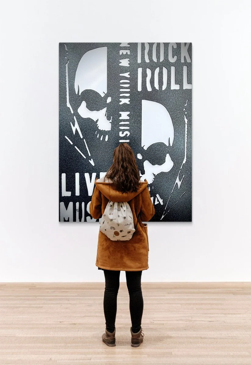 A woman with brown hair, wearing a brown coat and black pants, standing on a light wooden floor in an art gallery, observing a black and white artwork of two skulls and the words 'ROCK AND ROLL' and 'LIVE MUSIC' on a white wall.