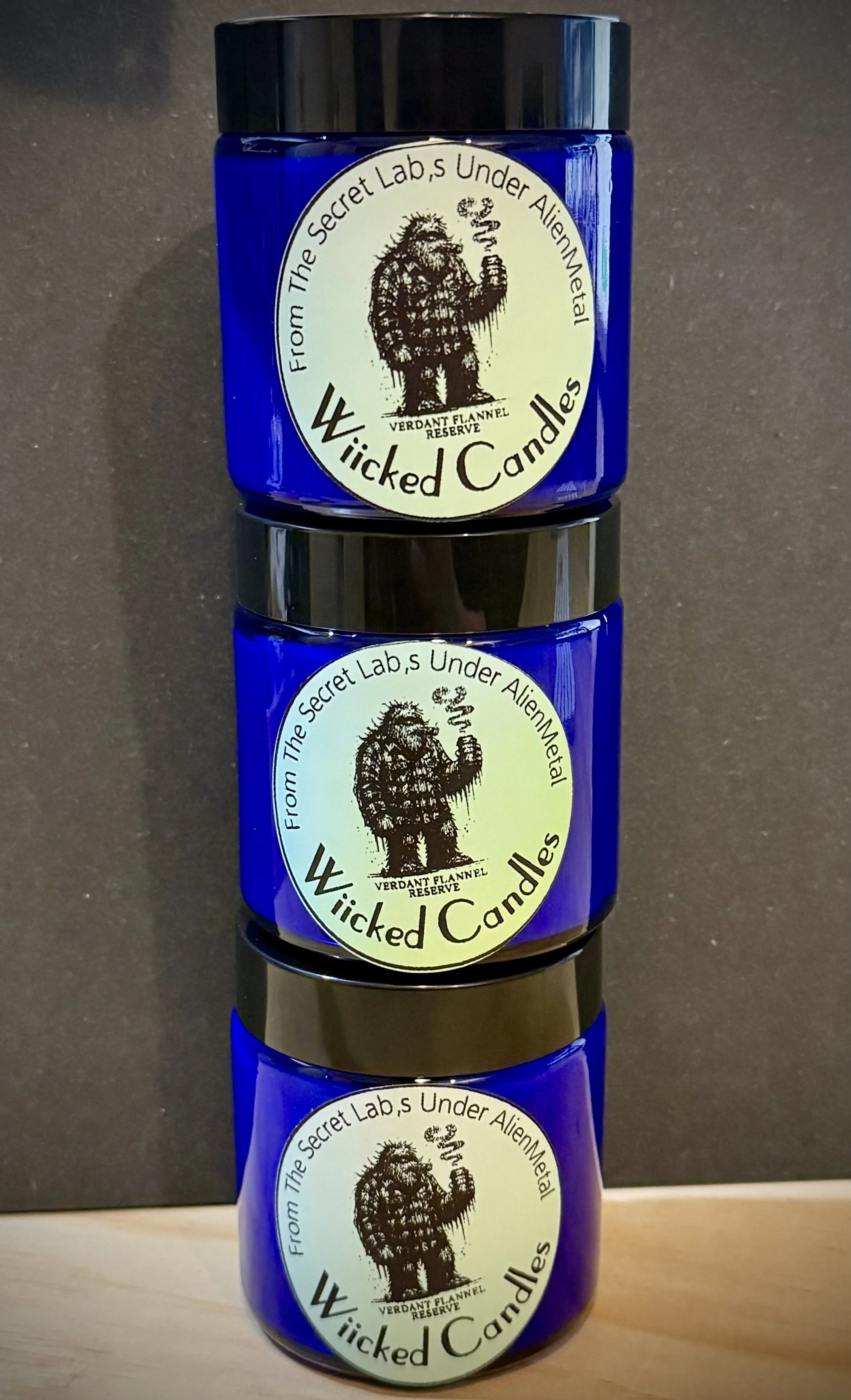 Three stacked blue candles with labels from 'Wicked Candles,' featuring a black and white illustration of a hairy creature holding a smoking object, surrounded by text.