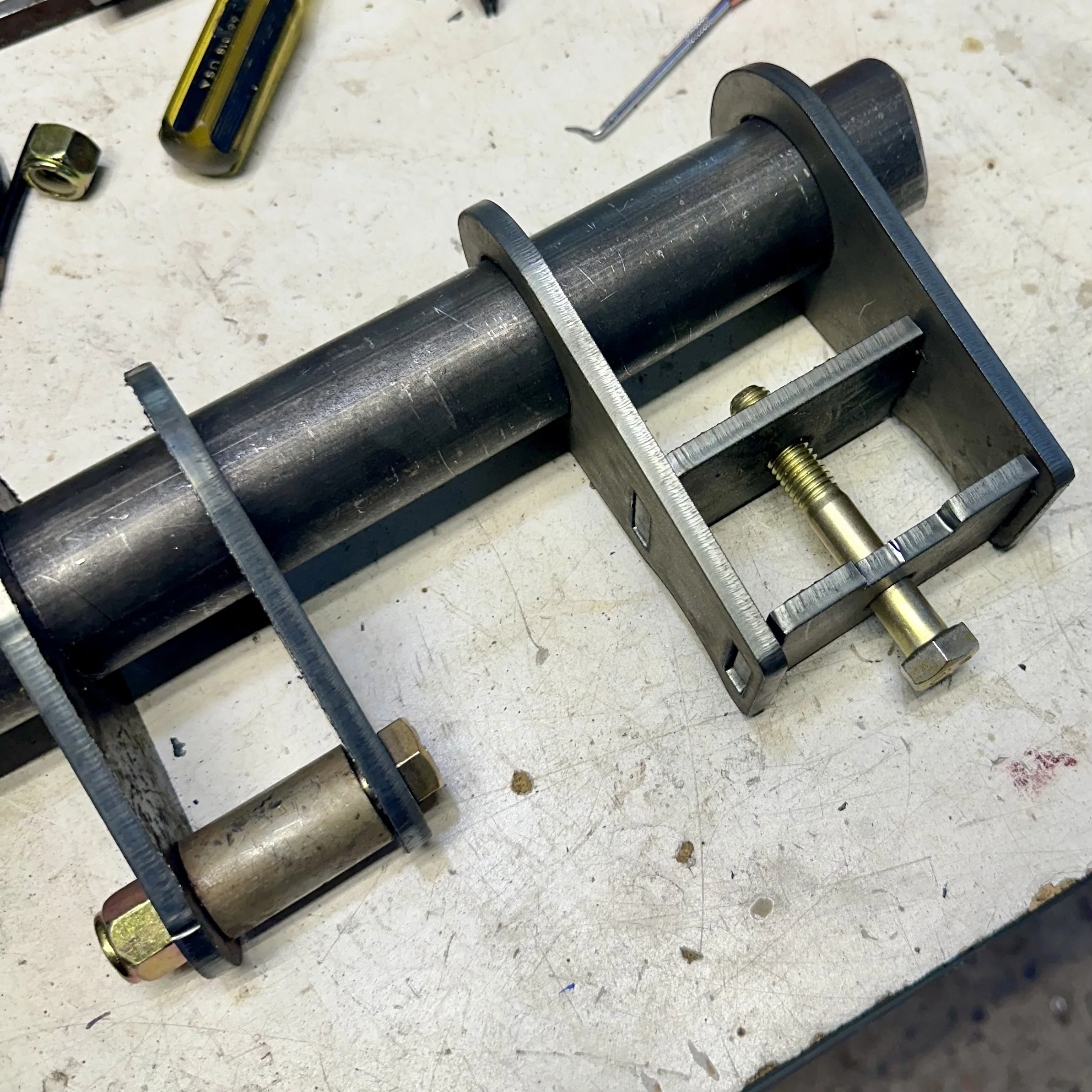 Close-up photograph of a mechanical component, possibly part of a machine or device, with a large metal shaft or rod secured by a metal frame with visible gold-colored bolts on a workbench or table with scattered tools and debris.