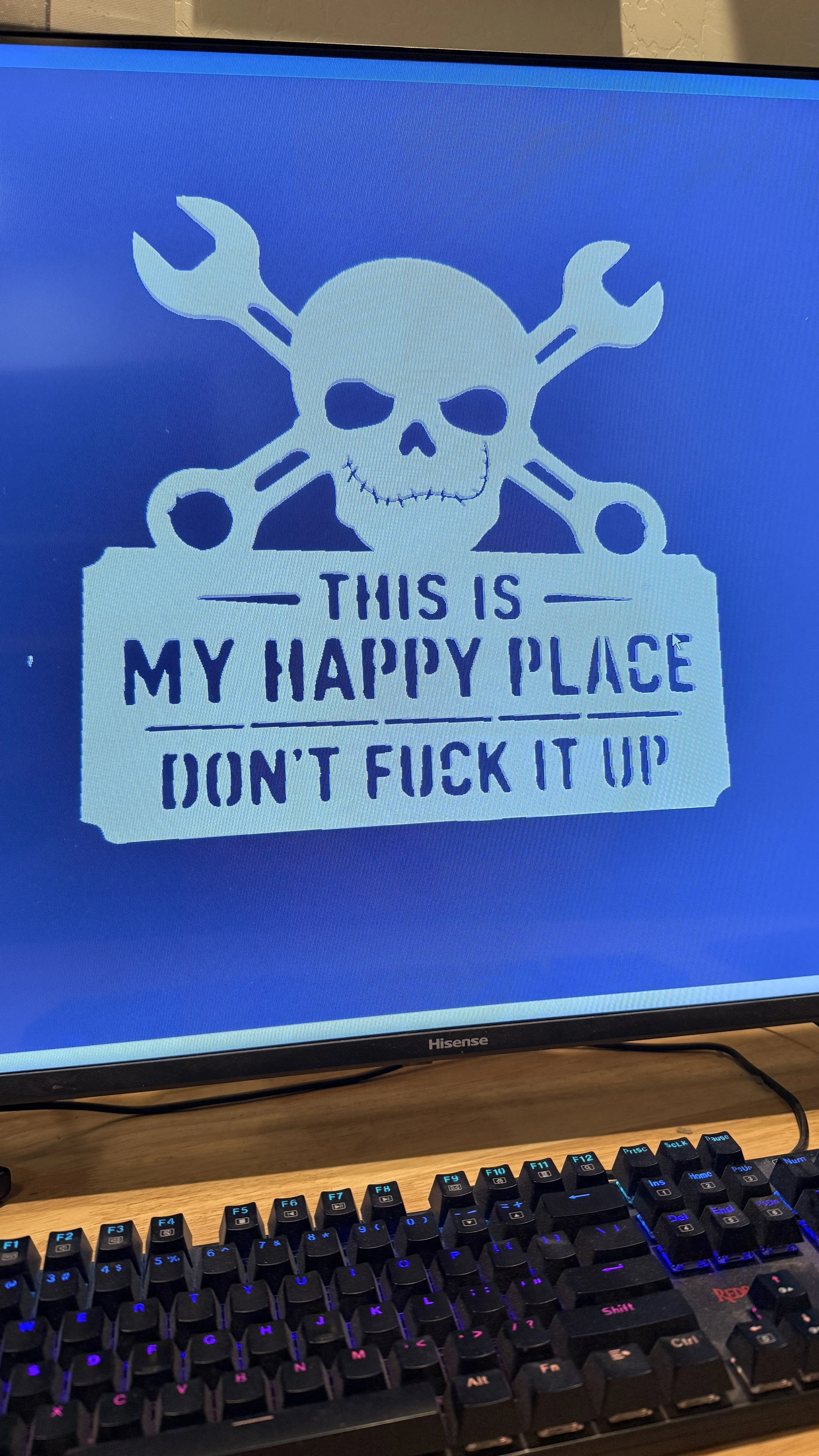 Computer monitor displays a skull with crossbones made of wrenches and a rebellious message that says, 'This is my happy place don't f*** it up', in a graffiti-style font.