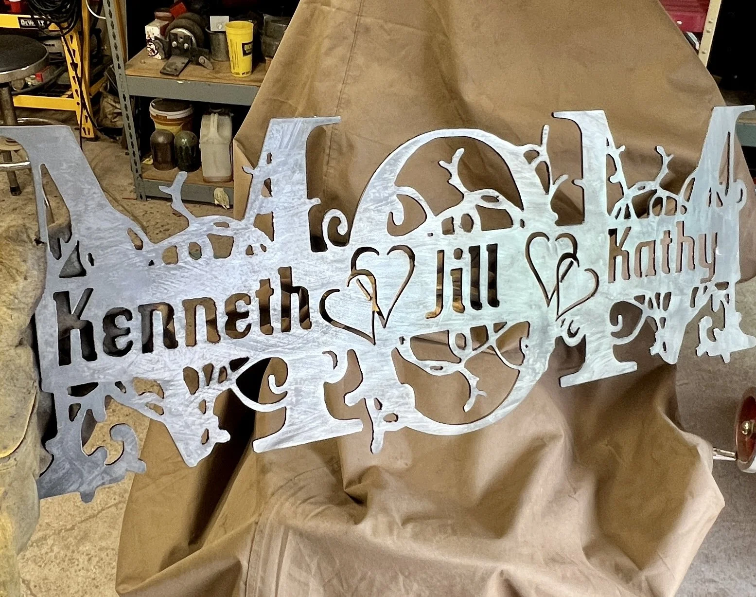 A decorative metal sign with intricate cutouts featuring the names 'Kenneth,' 'Jill,' and 'Kathy' with hearts and a Christmas tree design in the background.