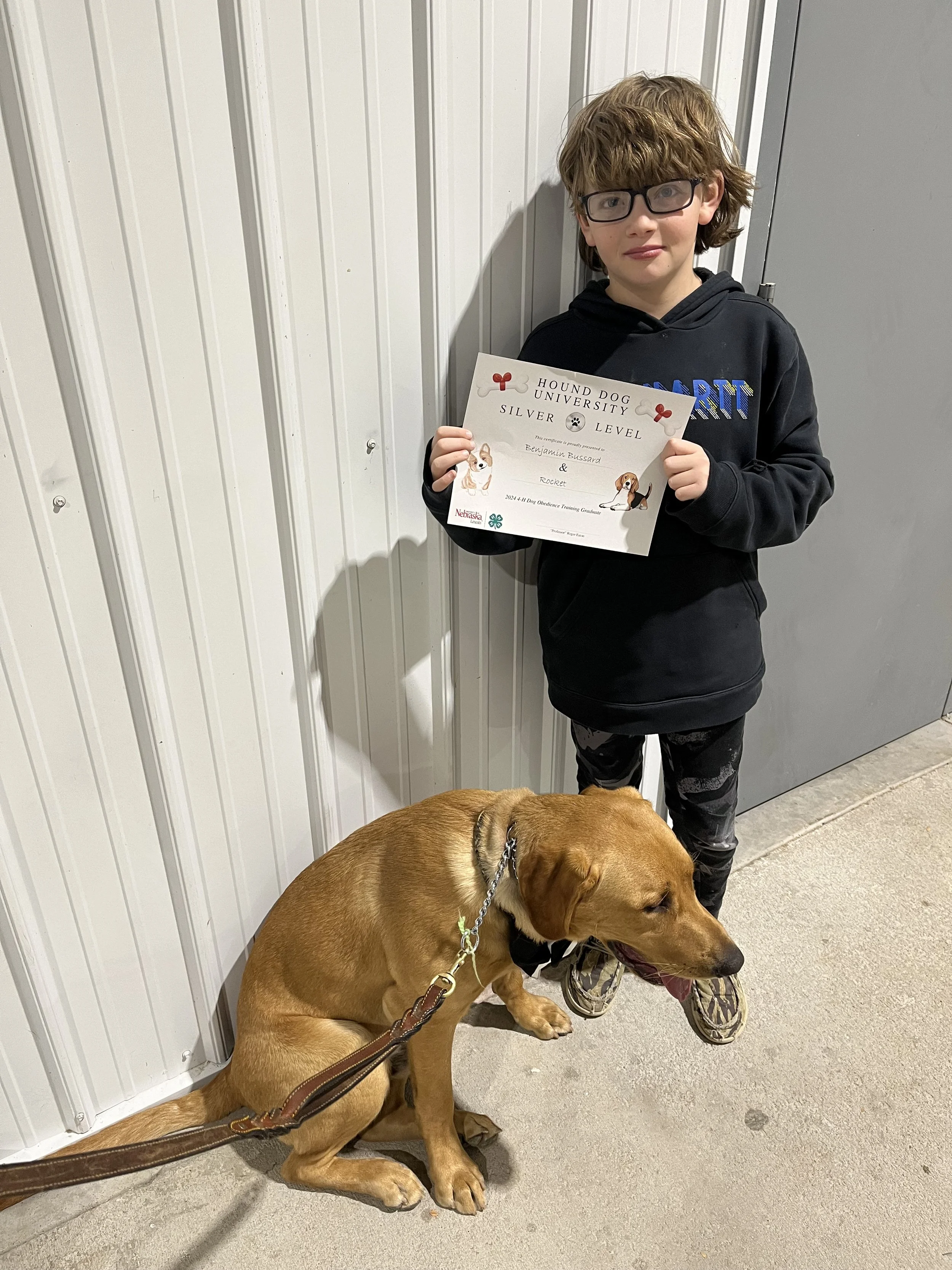 Young boy with glasses holding a certificate, standing next to a large brown dog sitting on the floor, against a white wall with vertical panels.