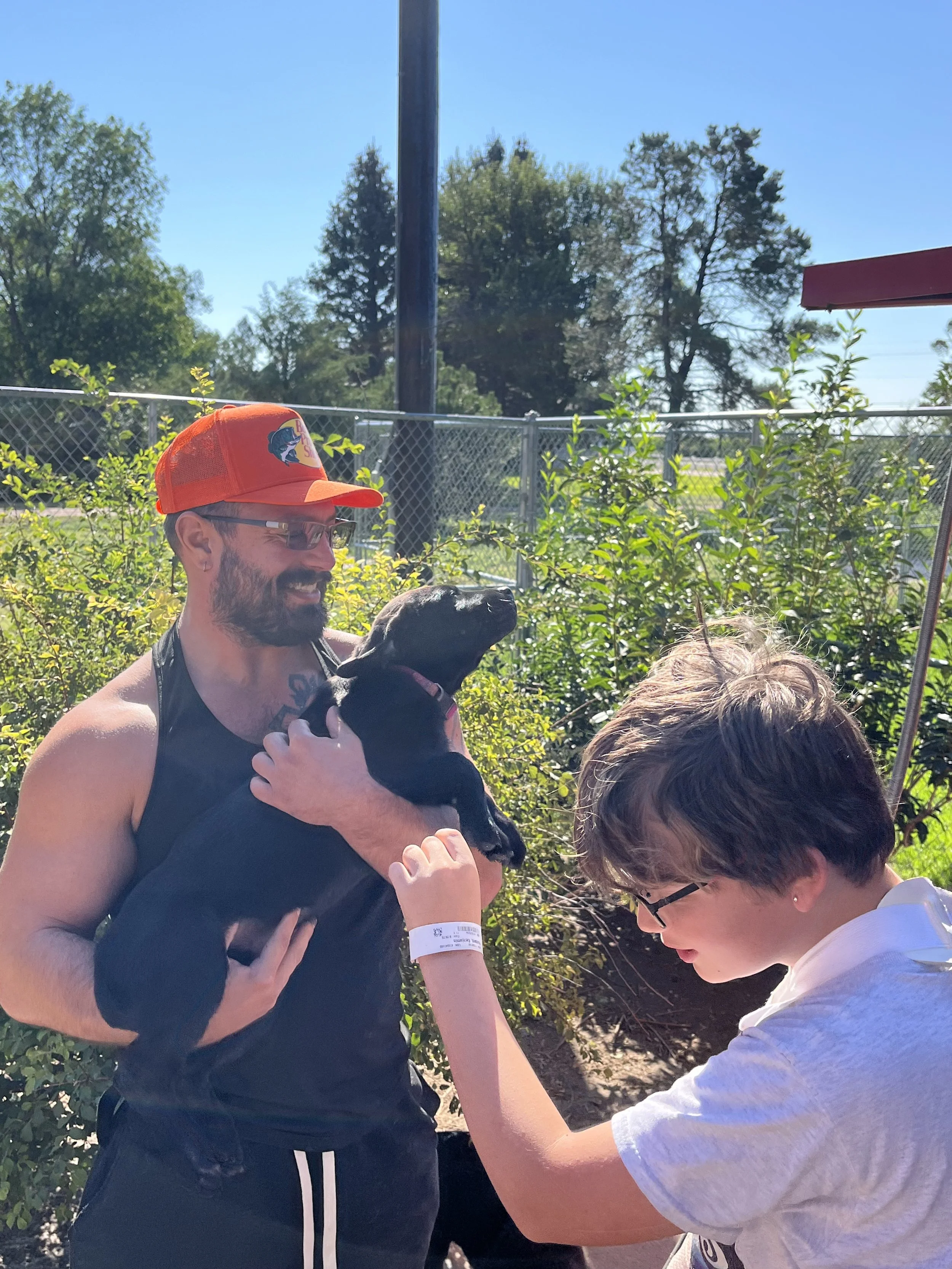 A young man with a beard and glasses, wearing a red cap and a black tank top, is holding a small black dog with his arm around it. Another person with short brown hair and a gray shirt is reaching out to the dog. They are outdoors on a sunny day, with trees, a chain-link fence, and a red structure visible in the background.