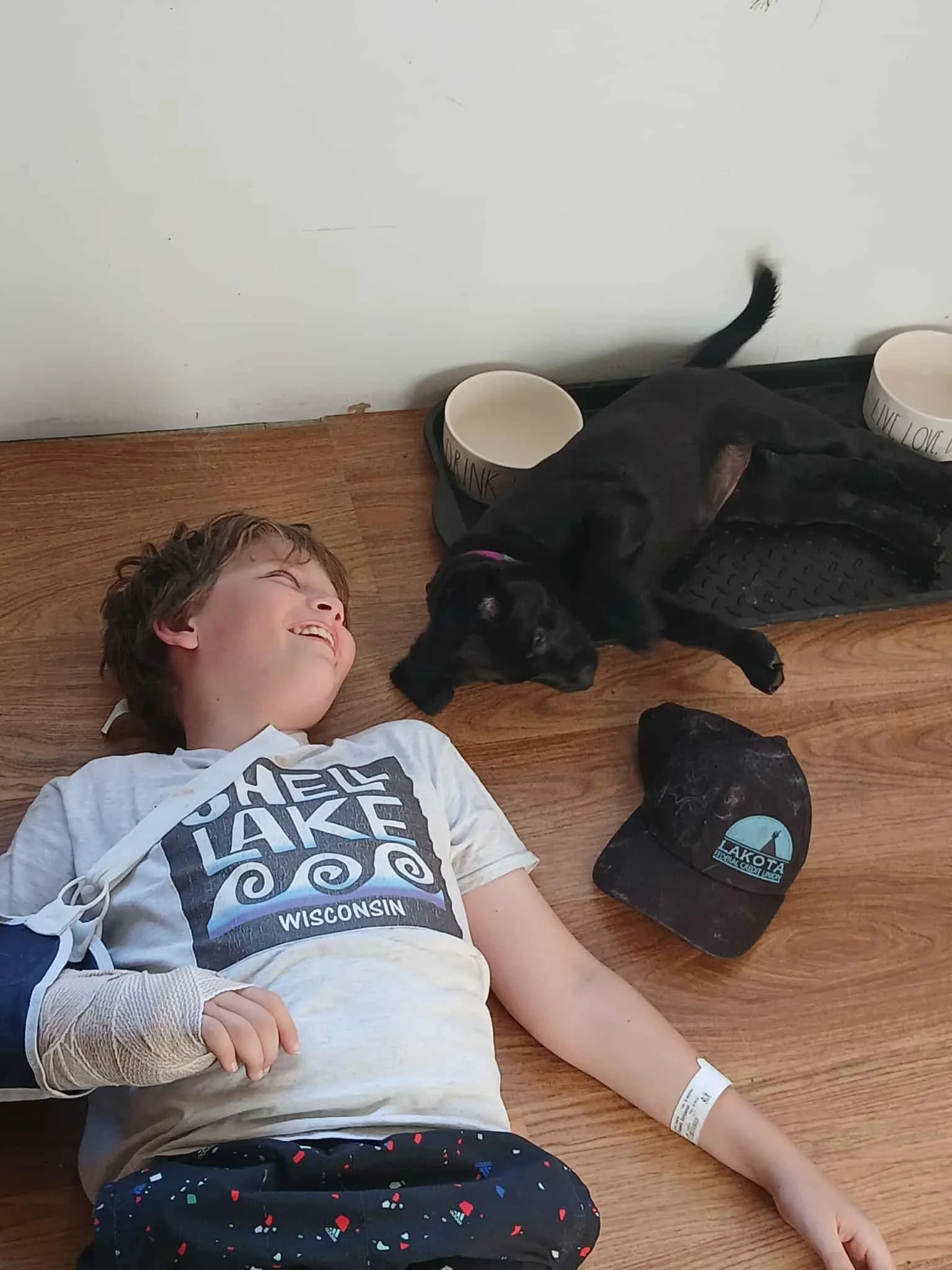 A young boy with a white T-shirt and dark shorts lies on a wooden floor, smiling, with a black puppy nursing from him. The puppy is near a black crate and two white bowls on a black mat. The boy has a hospital wristband on his wrist.