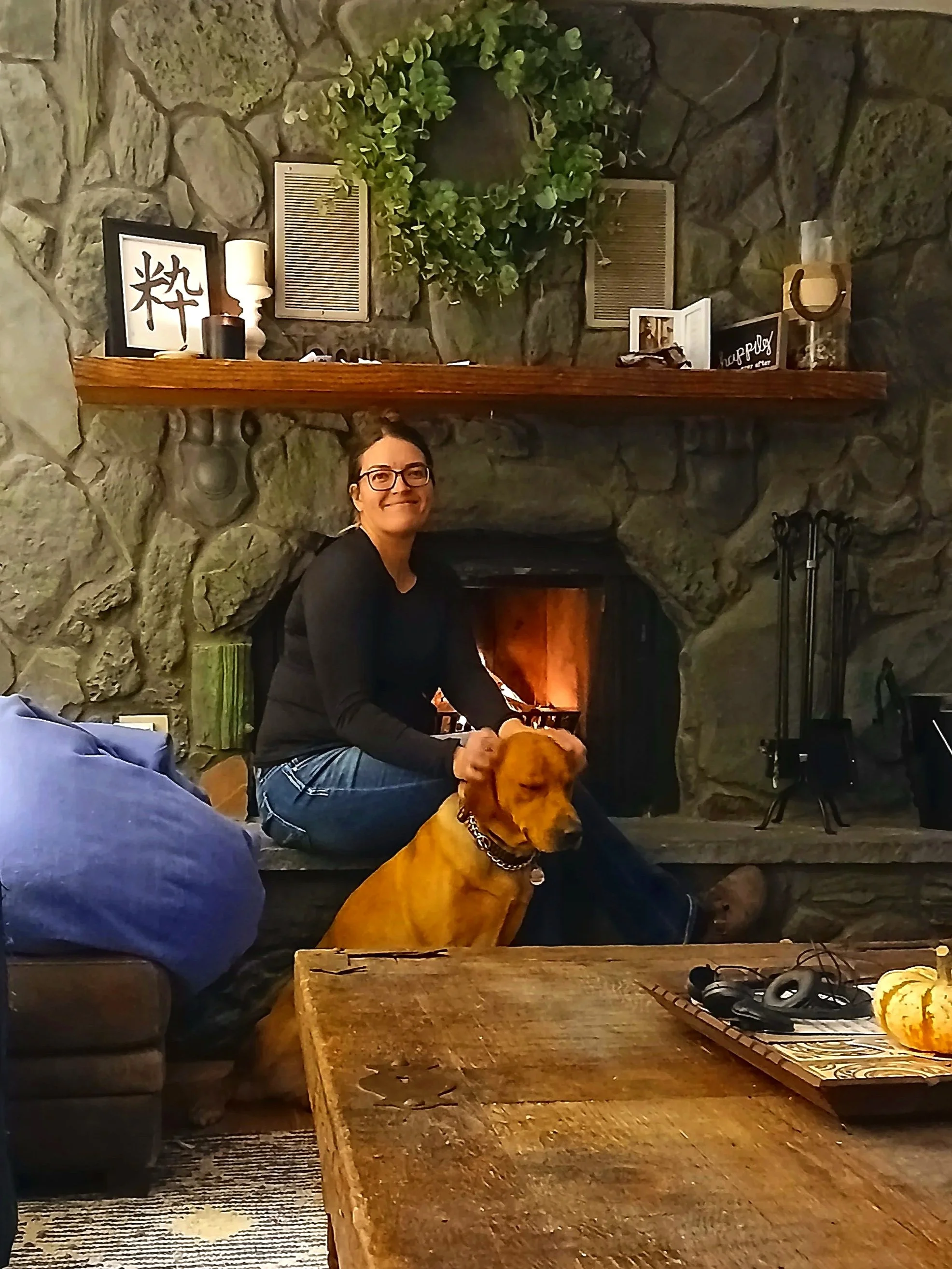 A woman with glasses smiling while sitting on a wooden bench in front of a stone fireplace, with a yellow dog sitting beside her. The mantel above the fireplace has a framed picture, some candles, a green plant, and decorative items. The room has rustic decor with a cozy ambiance.