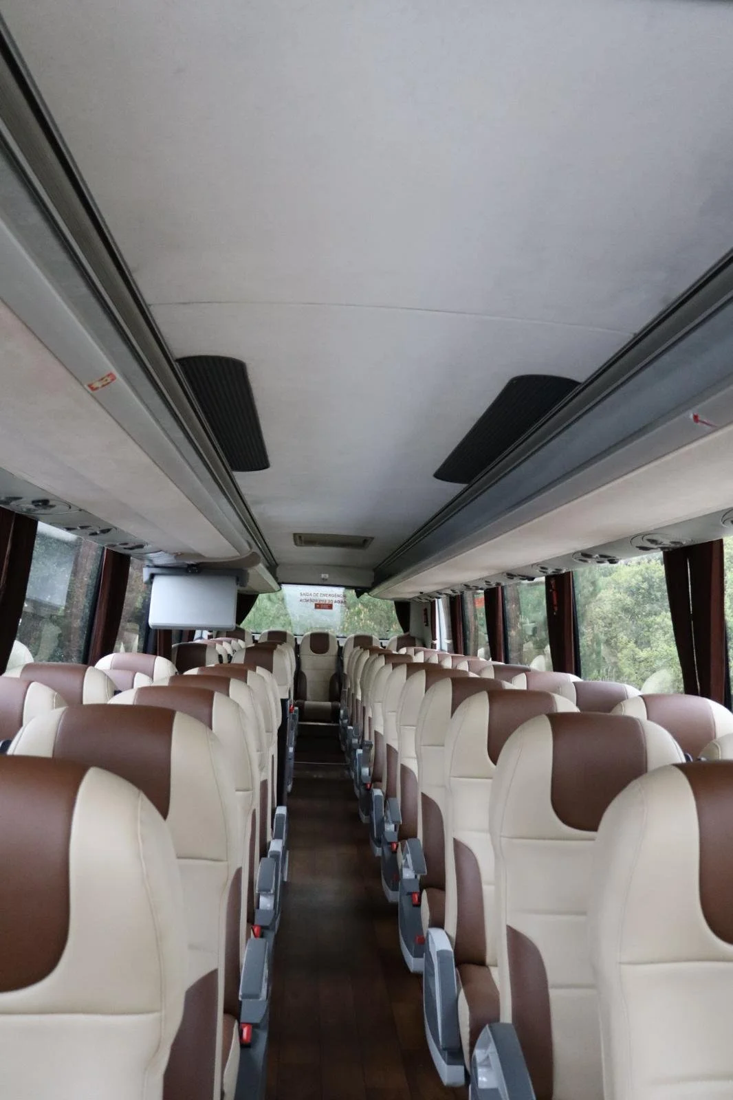 Interior de um ônibus com assentos em cores bege e marrom, janelas nas laterais e teto com iluminação e sistemas de ventilação.