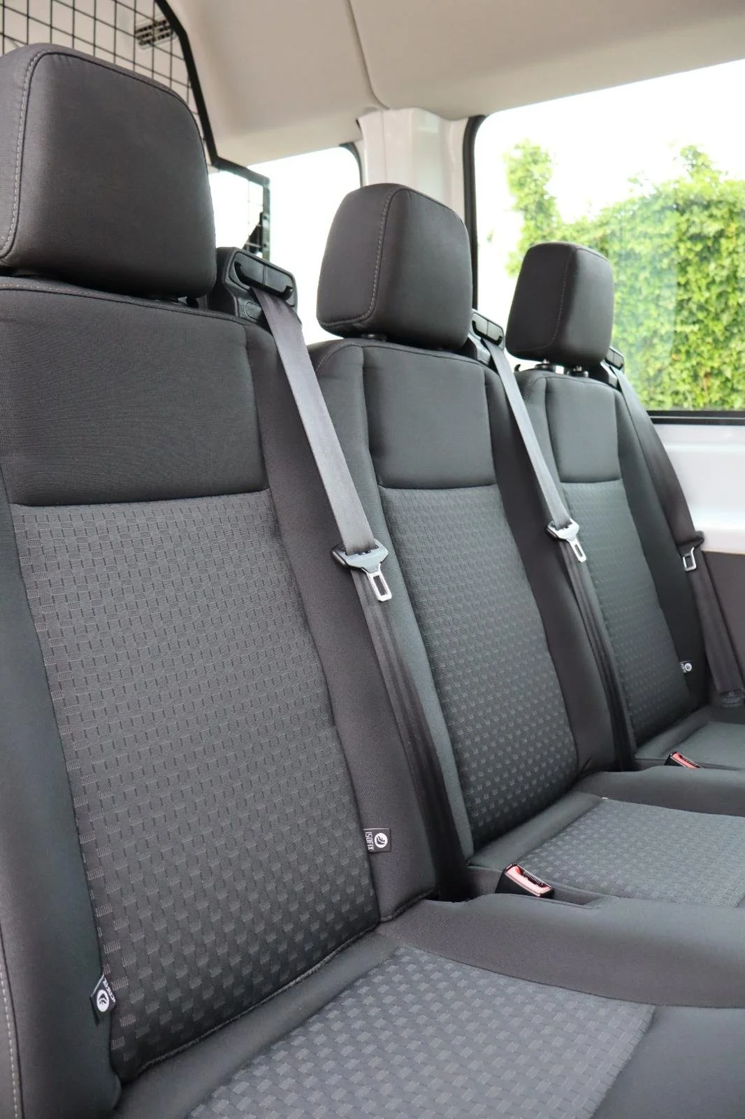 Assentos de carro com cintos de segurança e cabeceiras, janela ao lado com vista de árvores.
