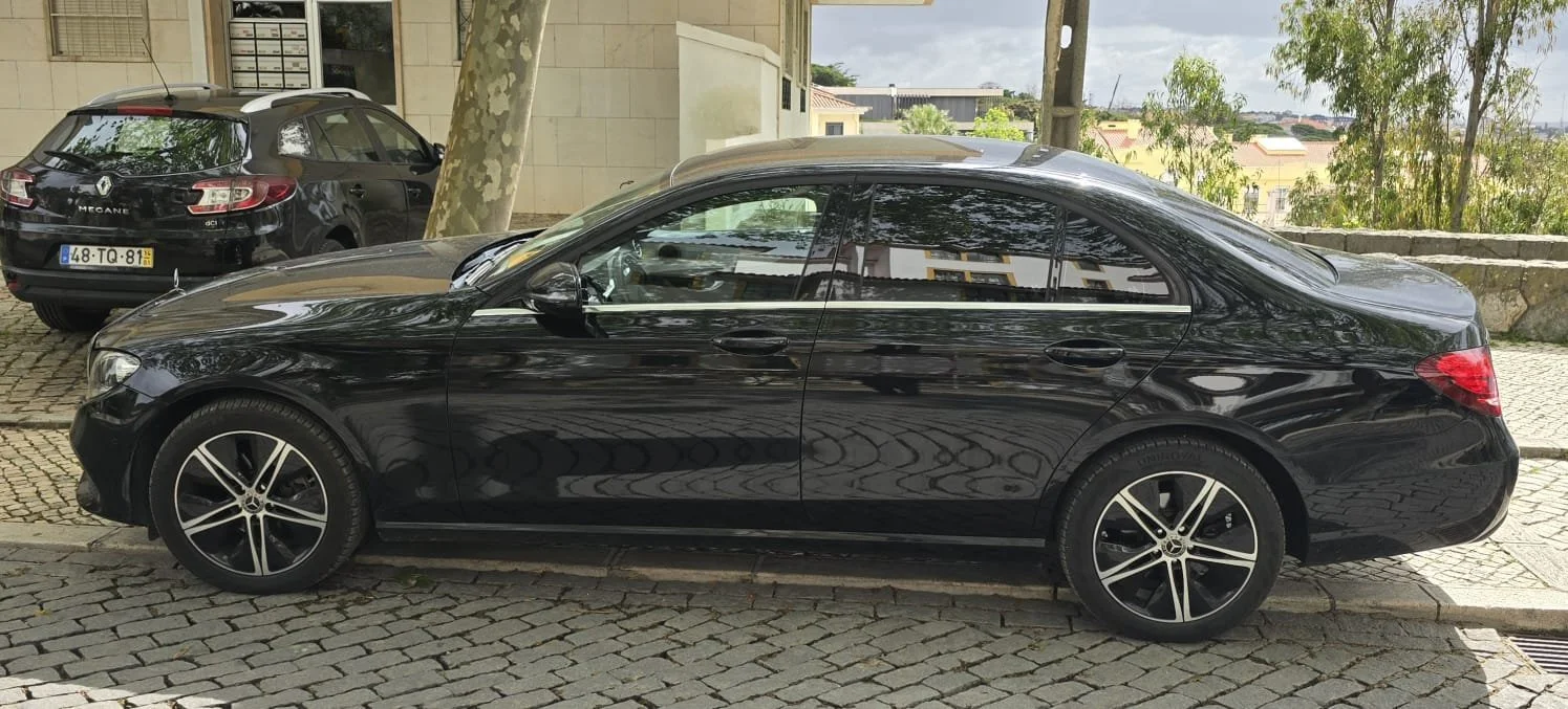 Carro preto estacionado na rua de paralelepípedos, com edifício ao fundo e árvores ao redor.