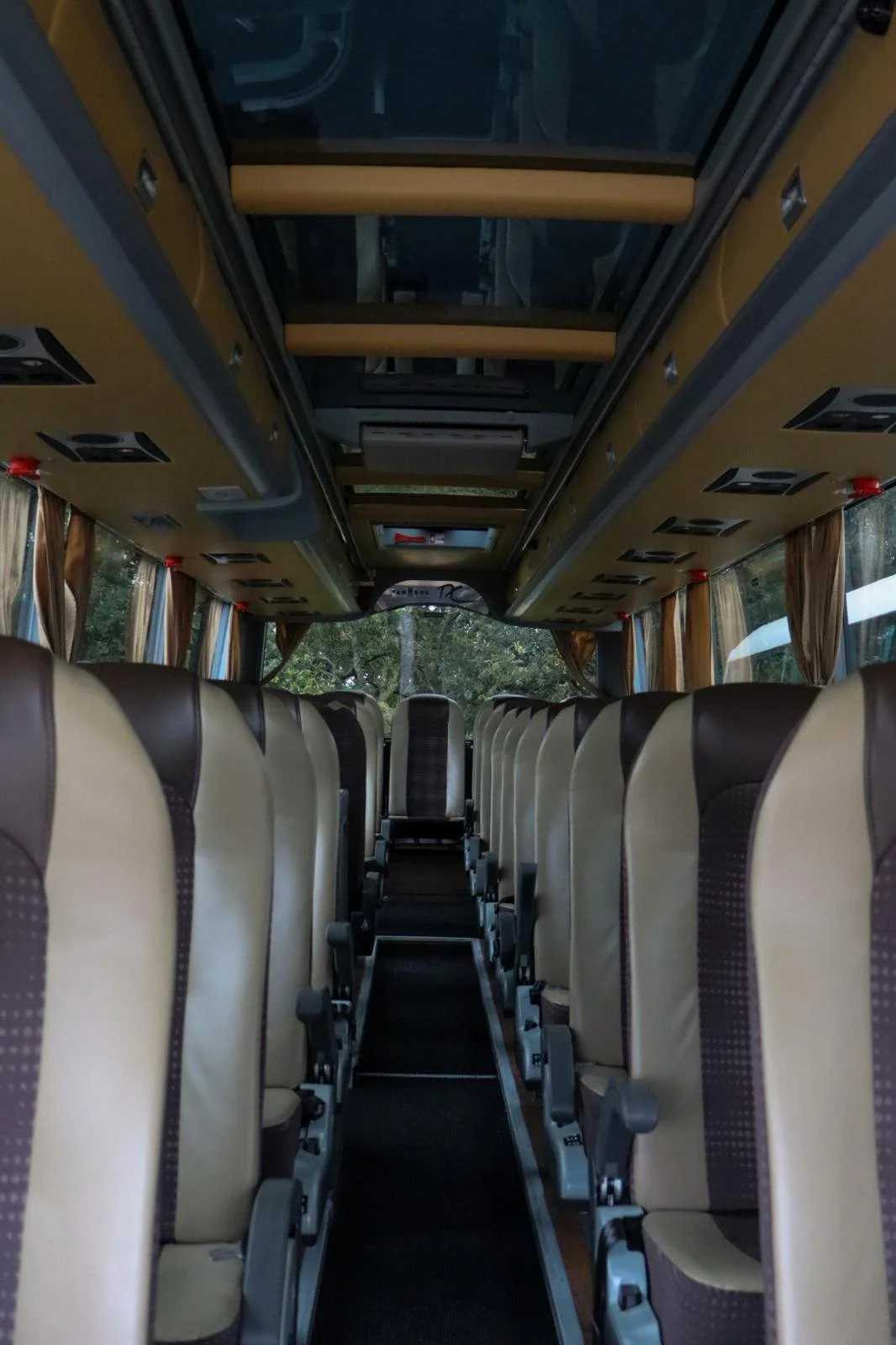 Interior de um ônibus com cadeiras alinhadas, cortinas nas janelas e teto equipado com luzes e compartimentos para bagagens.