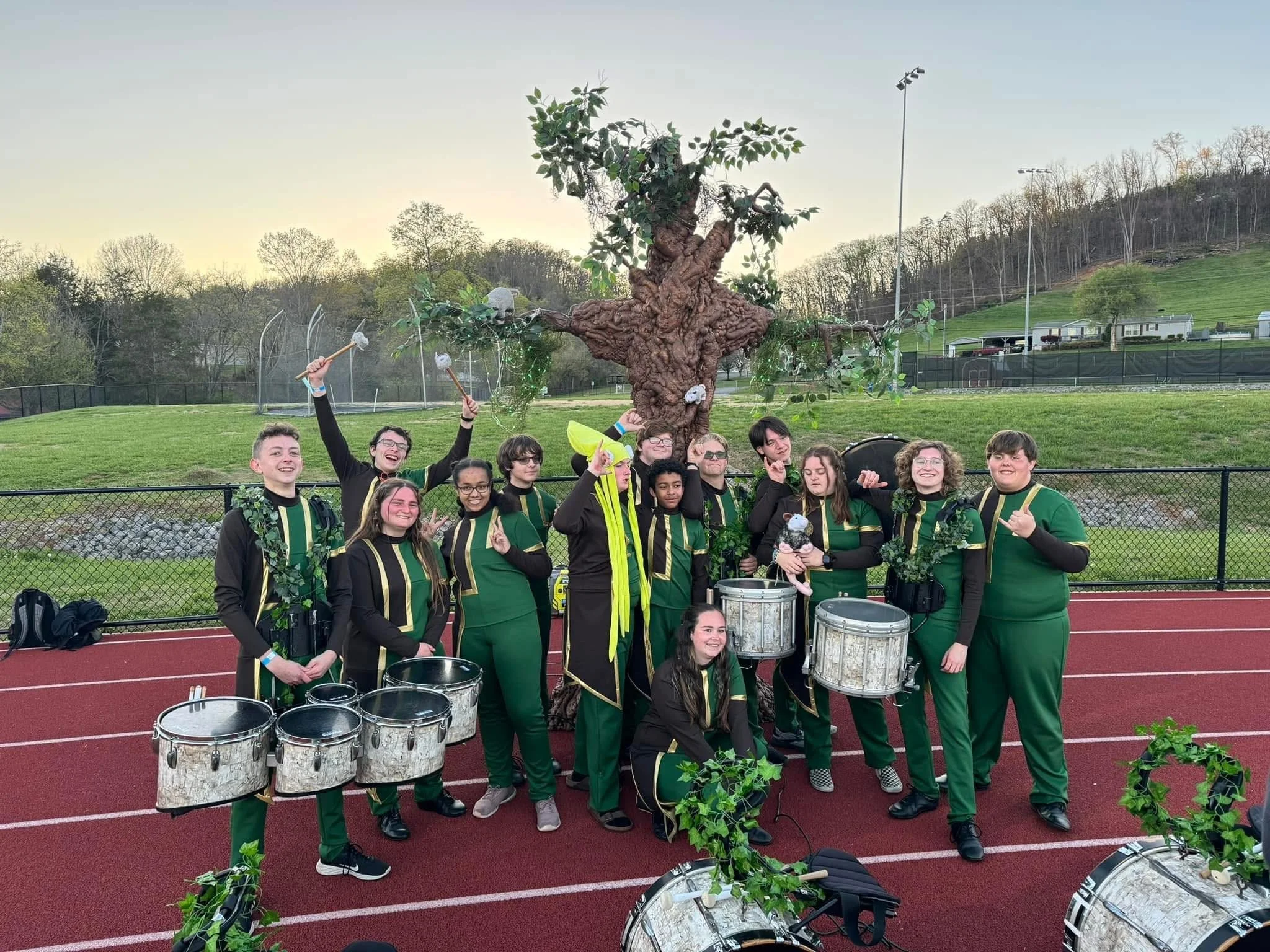 Group of children in band uniforms posing in front of a large, decorated tree on a sports field. The children are holding drums, drumsticks, and some are making peace signs, smiling and displaying excitement.