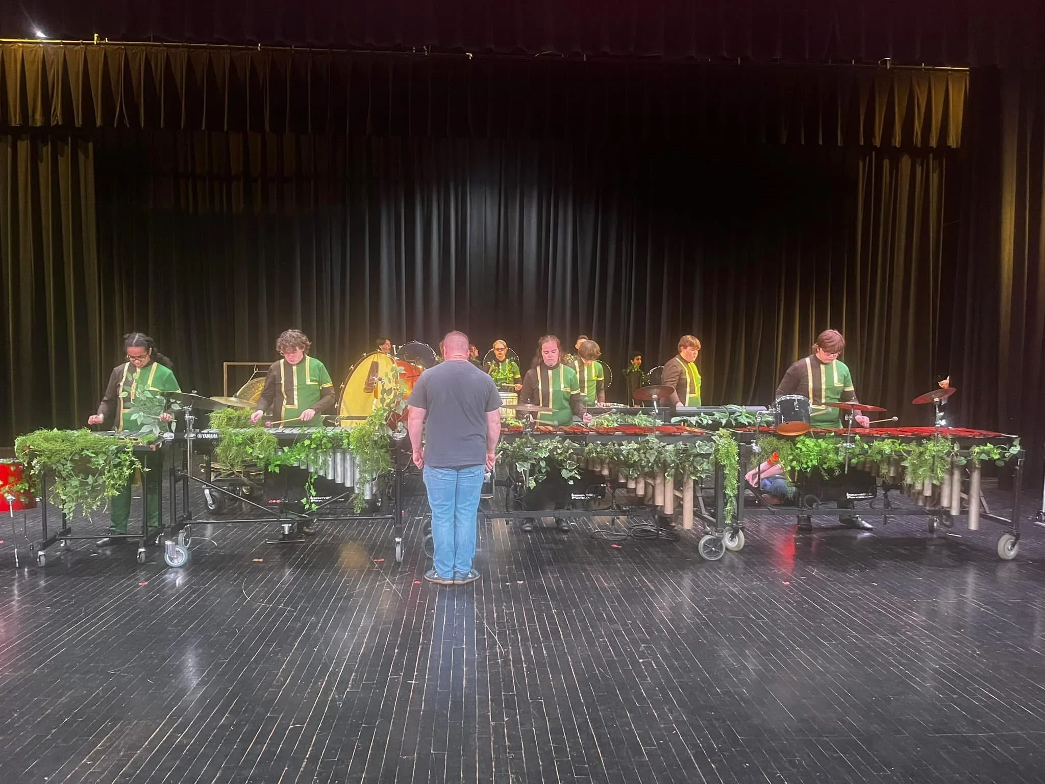 A group of musicians on stage, wearing green vests over black clothing, with various percussion instruments, in front of a large black curtain, with floral decorations on the front of the stage.