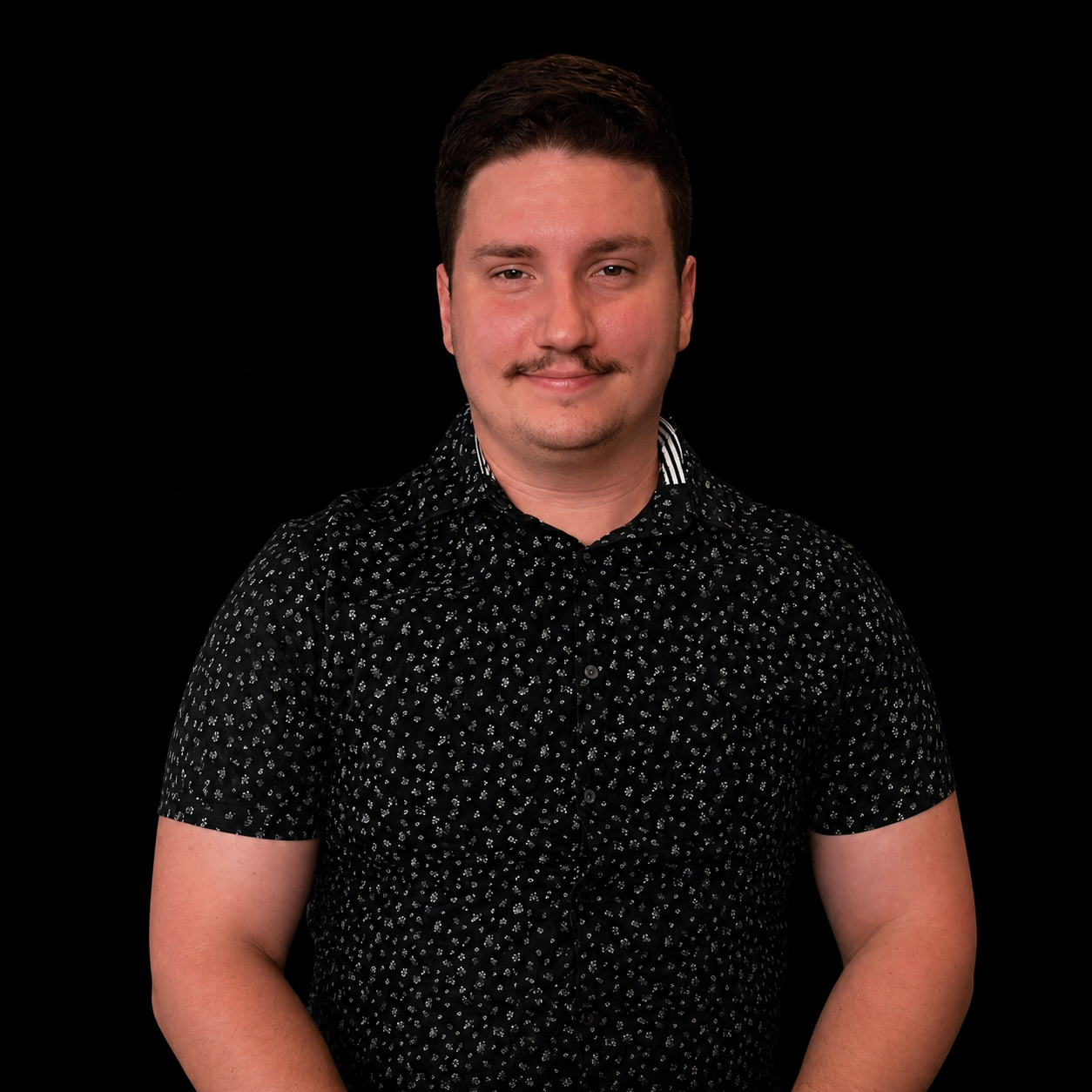A man with short dark hair, a mustache, and a slight smile, wearing a black shirt with a white floral pattern, standing against a black background.