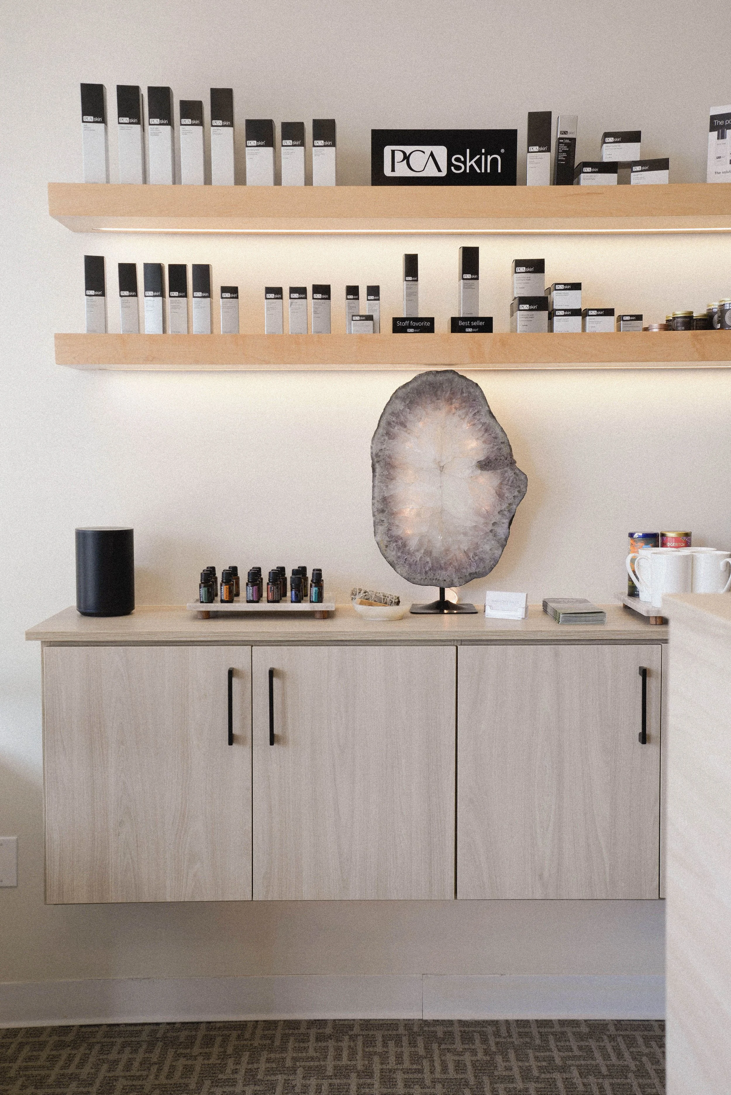 A wooden cabinet with black handles, mounted on a wall, with a large geode on top of it. Above the cabinet are two wooden shelves holding PCA Skin products, with labels indicating a staff favorite and best seller. To the left of the cabinet is a black cylindrical speaker, and to the right are small bottles and cups on a small tray.
