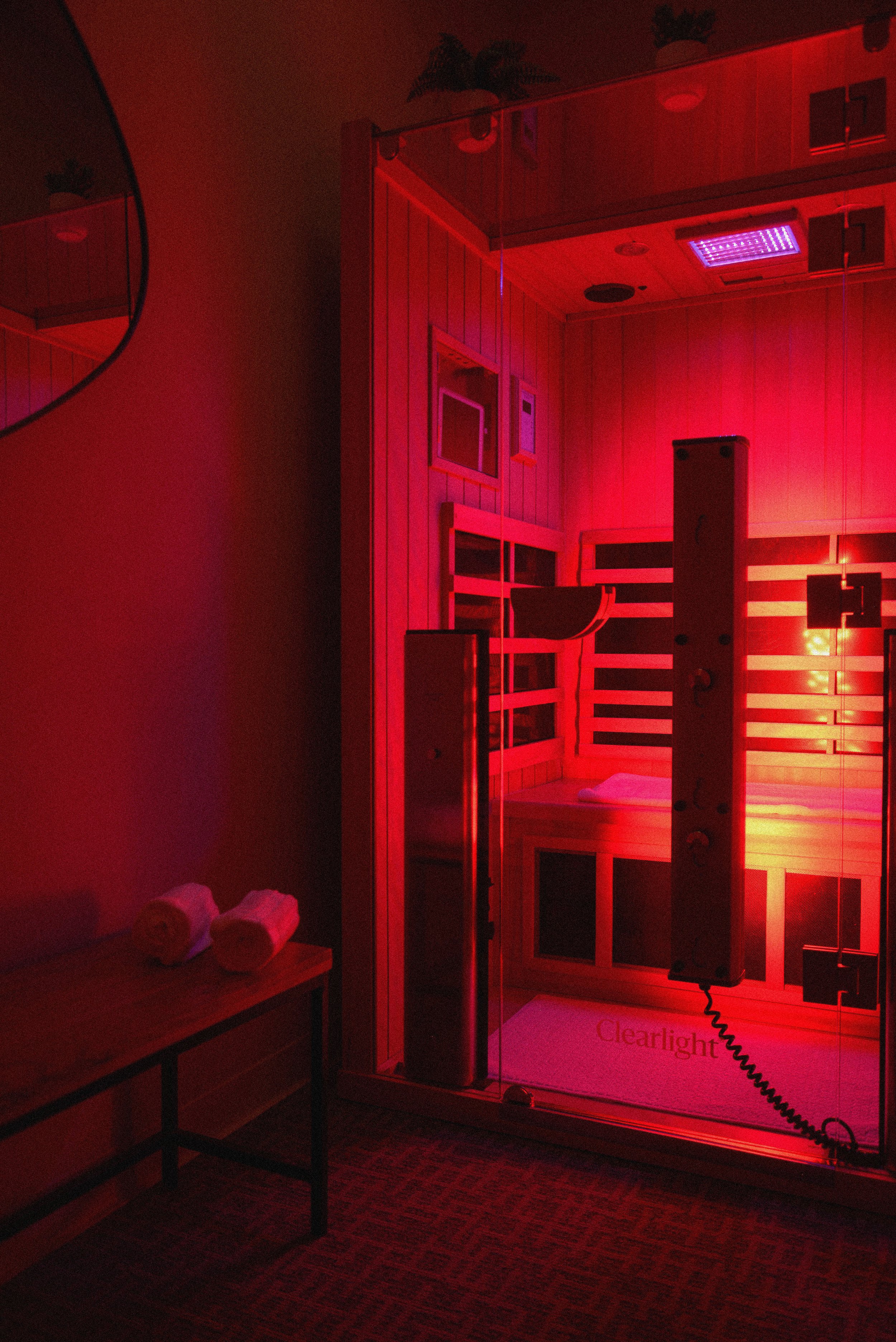 An infrared light therapy device, branded as 'Clearlight,' is positioned in a room with wood-paneled walls and a suite of furniture. The device emits a vivid red light, illuminating the surrounding area.