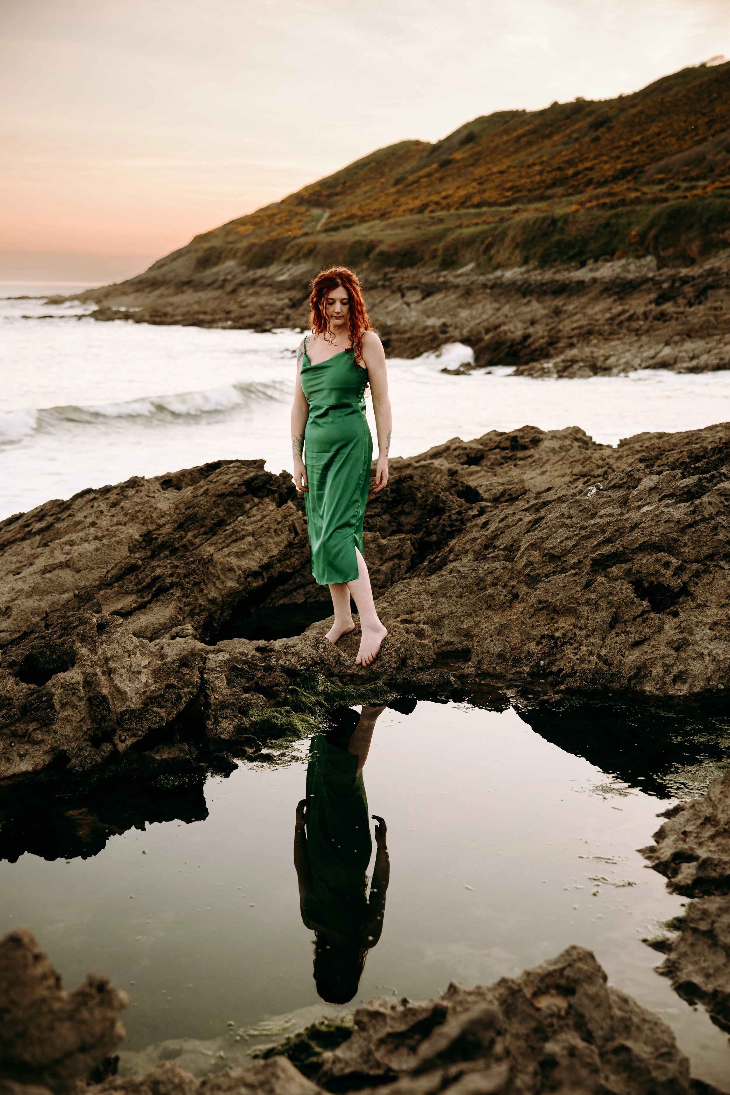 A woman in a green dress standing on rocks by the ocean, with her reflection visible in a small pool of water, at sunset or dusk.
