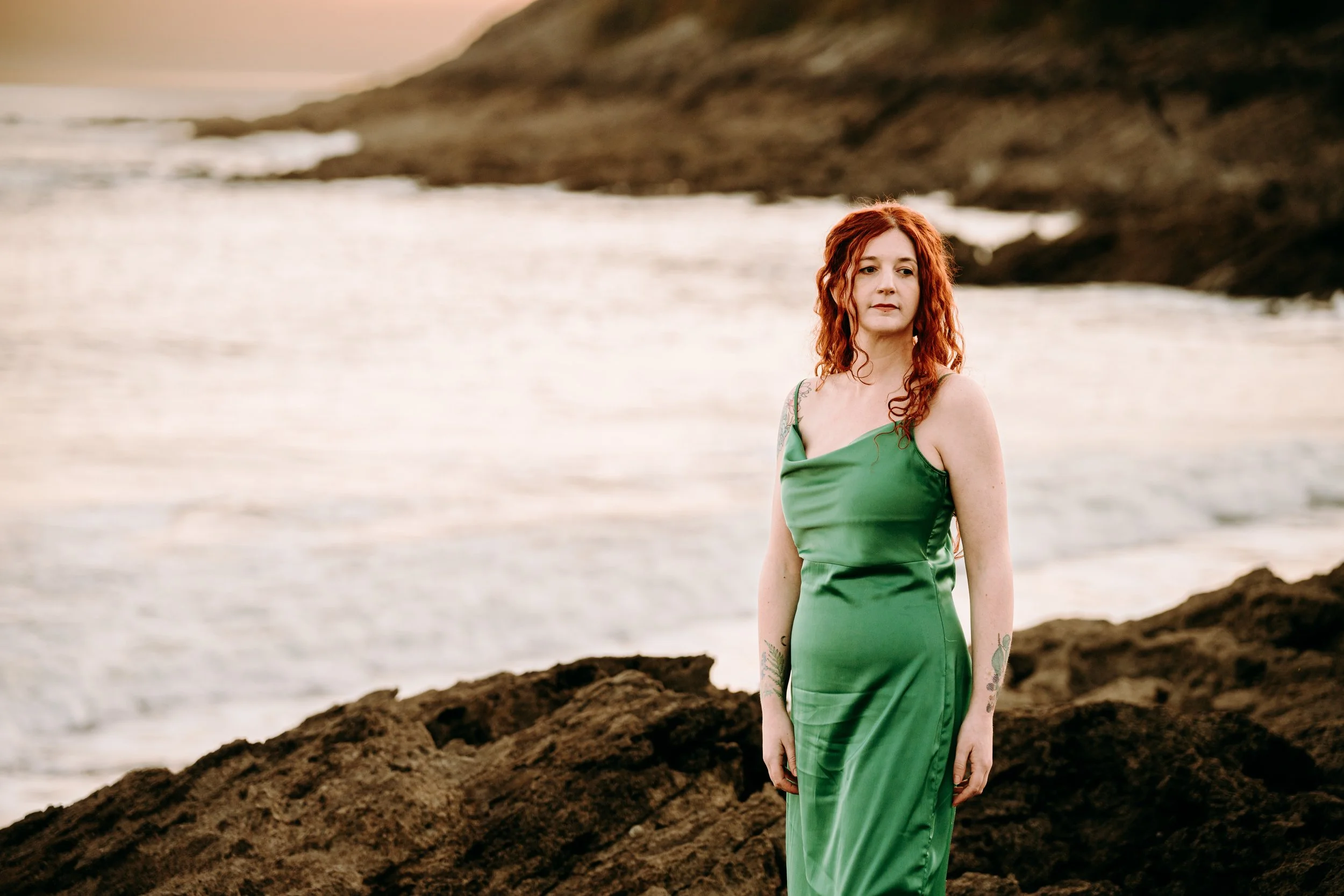 Hermione Wild, a woman in folk music, with red hair and tattoos wearing a green dress standing on a rocky shoreline with the ocean and distant cliffs in the background at sunset.