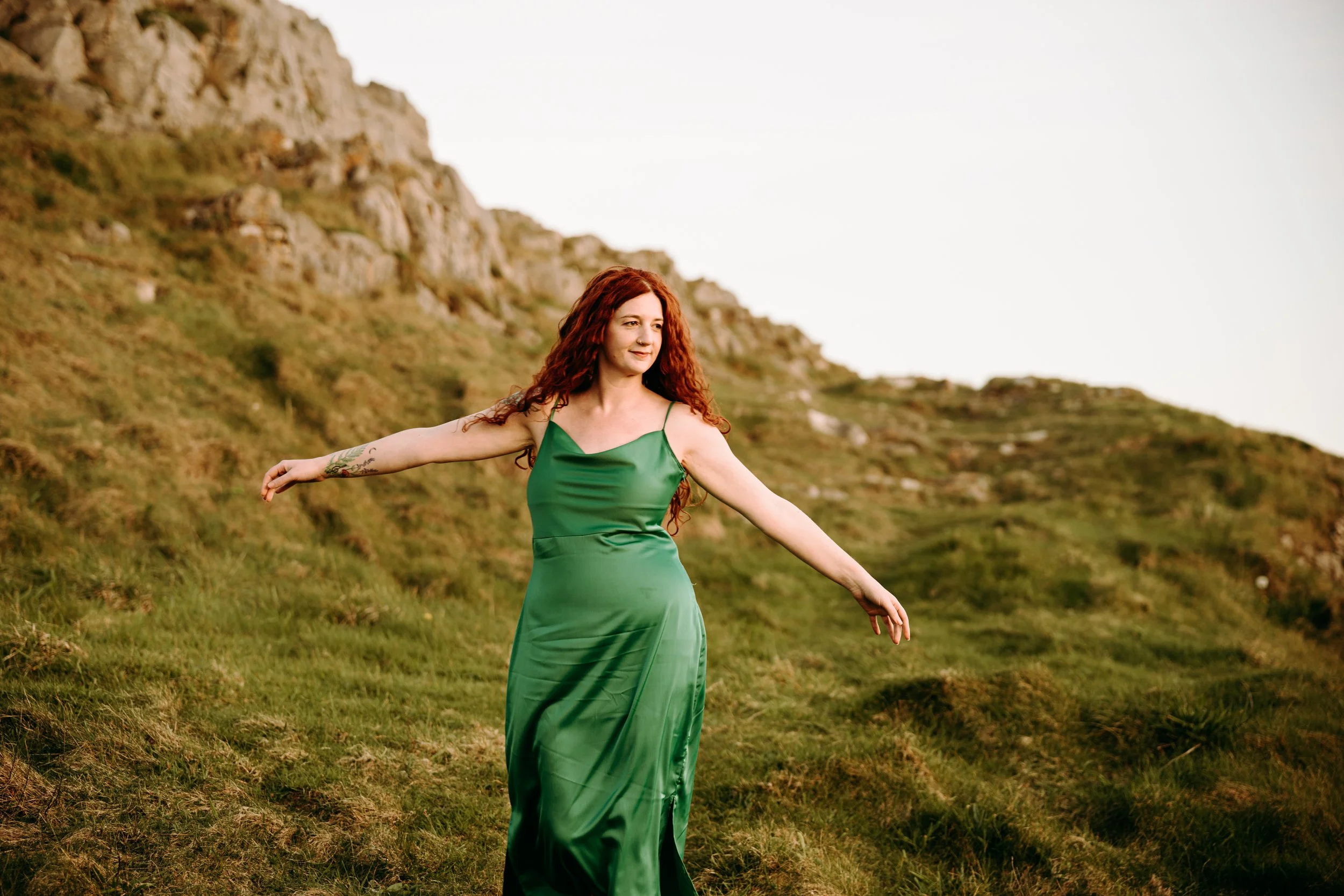Hermione Wild, a female folk musician with long red hair wearing a green satin dress standing outdoors on a hillside with grass and rocky cliffs in the background, arms outstretched.