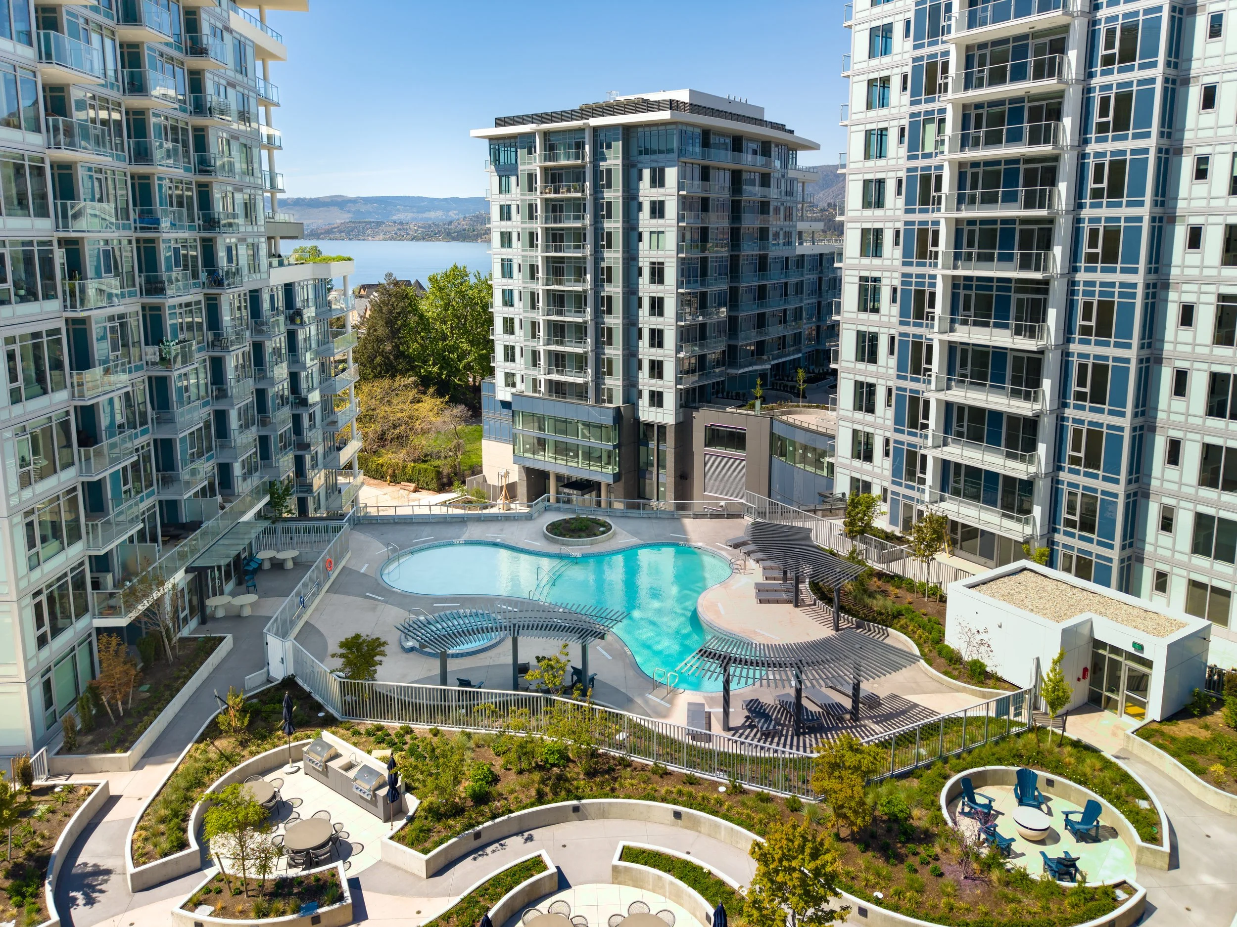 Modern apartment complex with glass-fronted buildings, a central swimming pool, outdoor seating, and scenic water and mountains in the background.