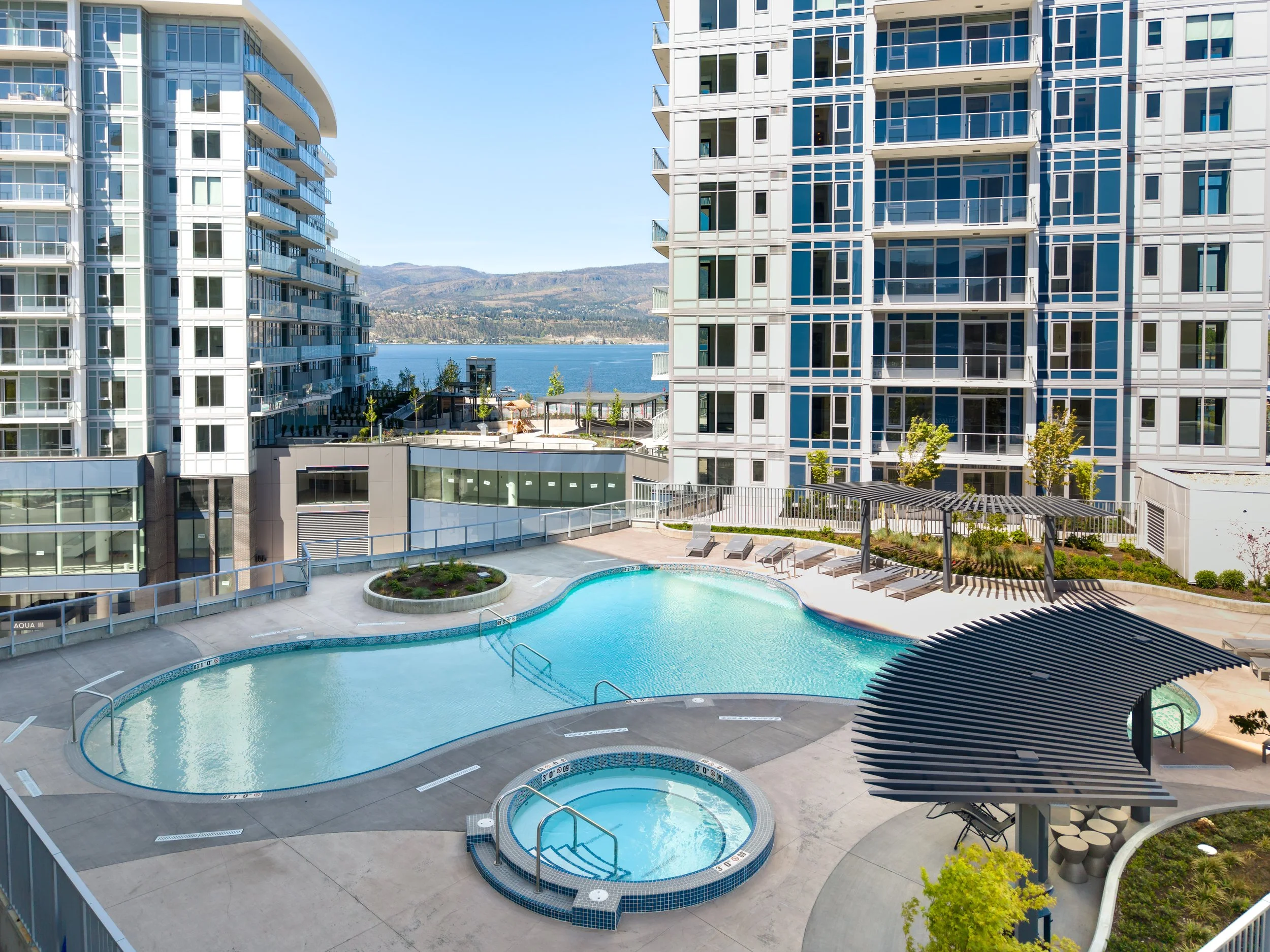 View of a modern apartment complex with a swimming pool area, lounge chairs, and a scenic backdrop of mountains and a body of water.
