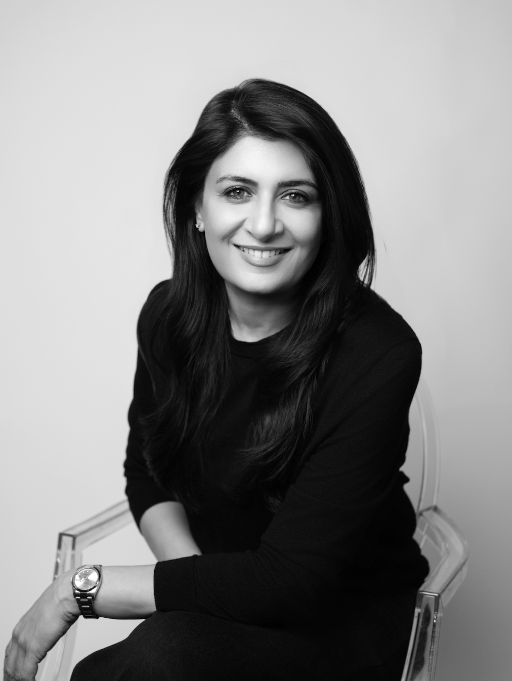 Black and white portrait of a smiling woman with long dark hair, sitting on a transparent chair, wearing a black top and a wristwatch.