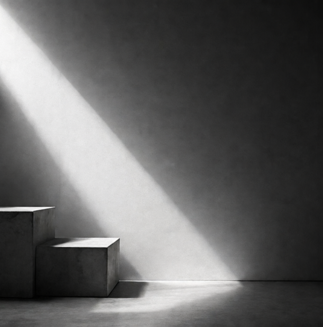 Black and white image of three concrete steps casting shadows on a bare wall and floor, with a beam of sunlight creating a diagonal light pattern.