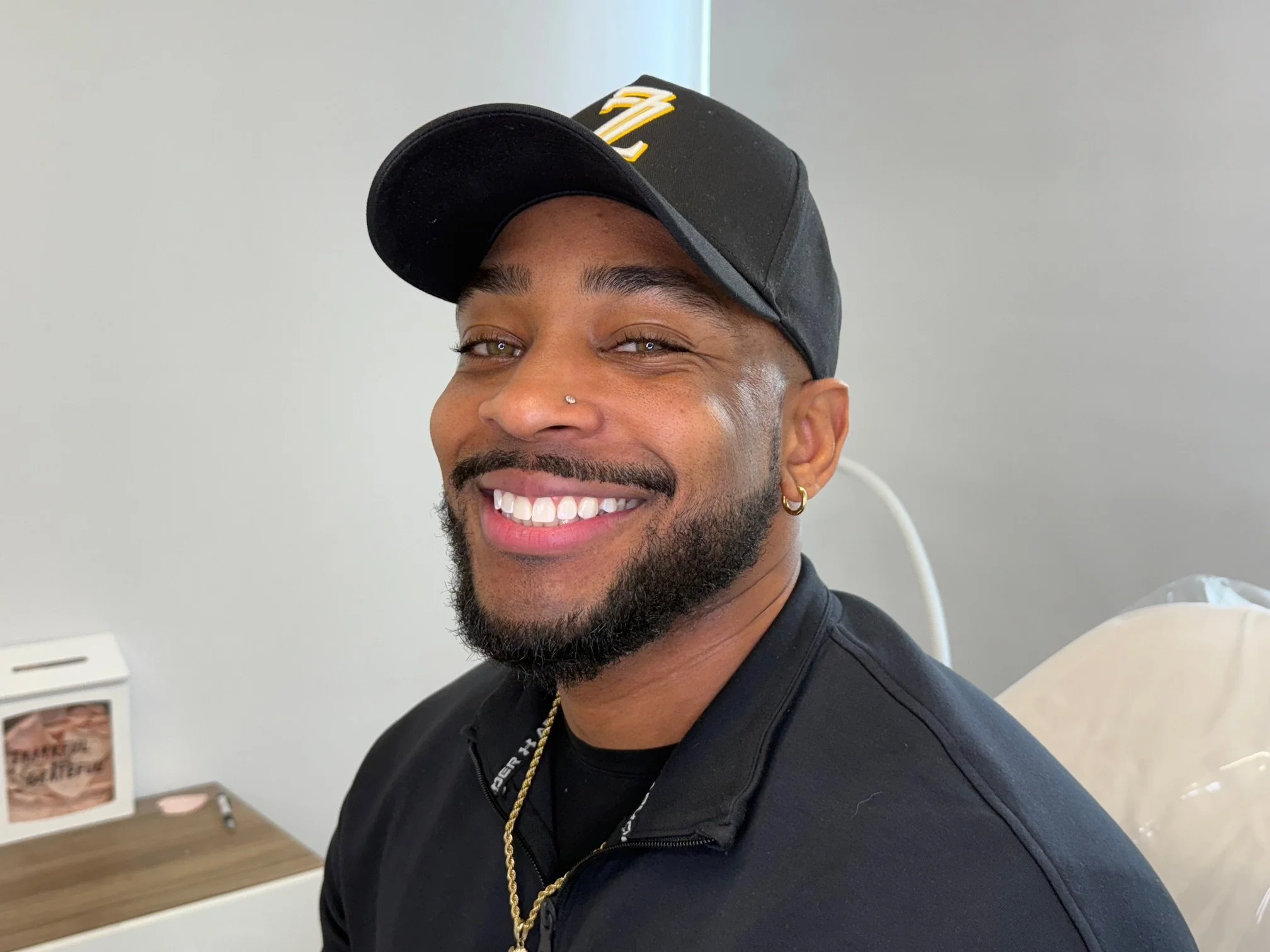 Close-up of a smiling man wearing a black cap with yellow and white logo, black jacket, and gold hoop earring, in a clinical setting.