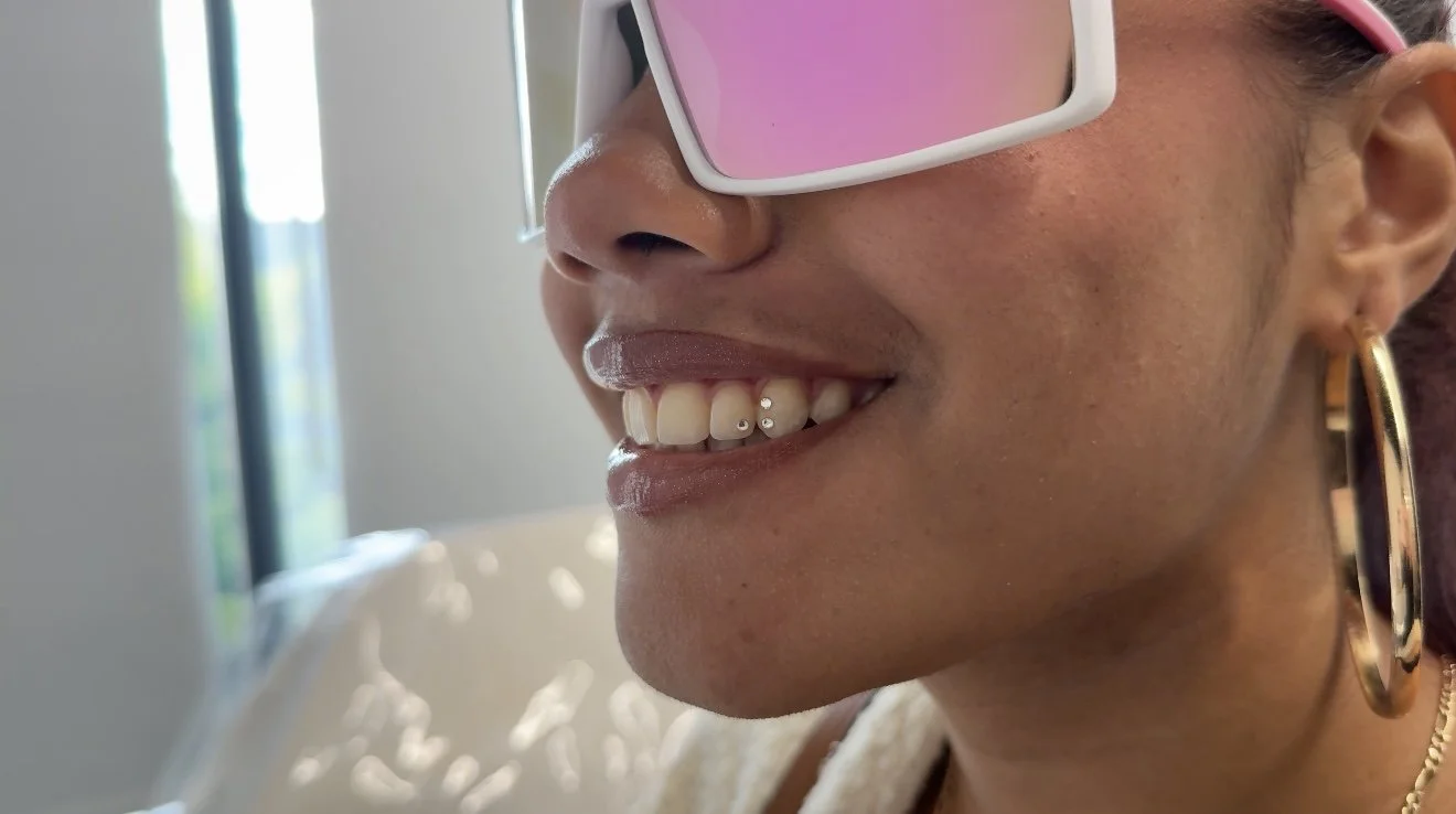 Close-up of a smiling woman with light brown skin, wearing white and pink sunglasses, gold hoop earrings, and a gold chain necklace.