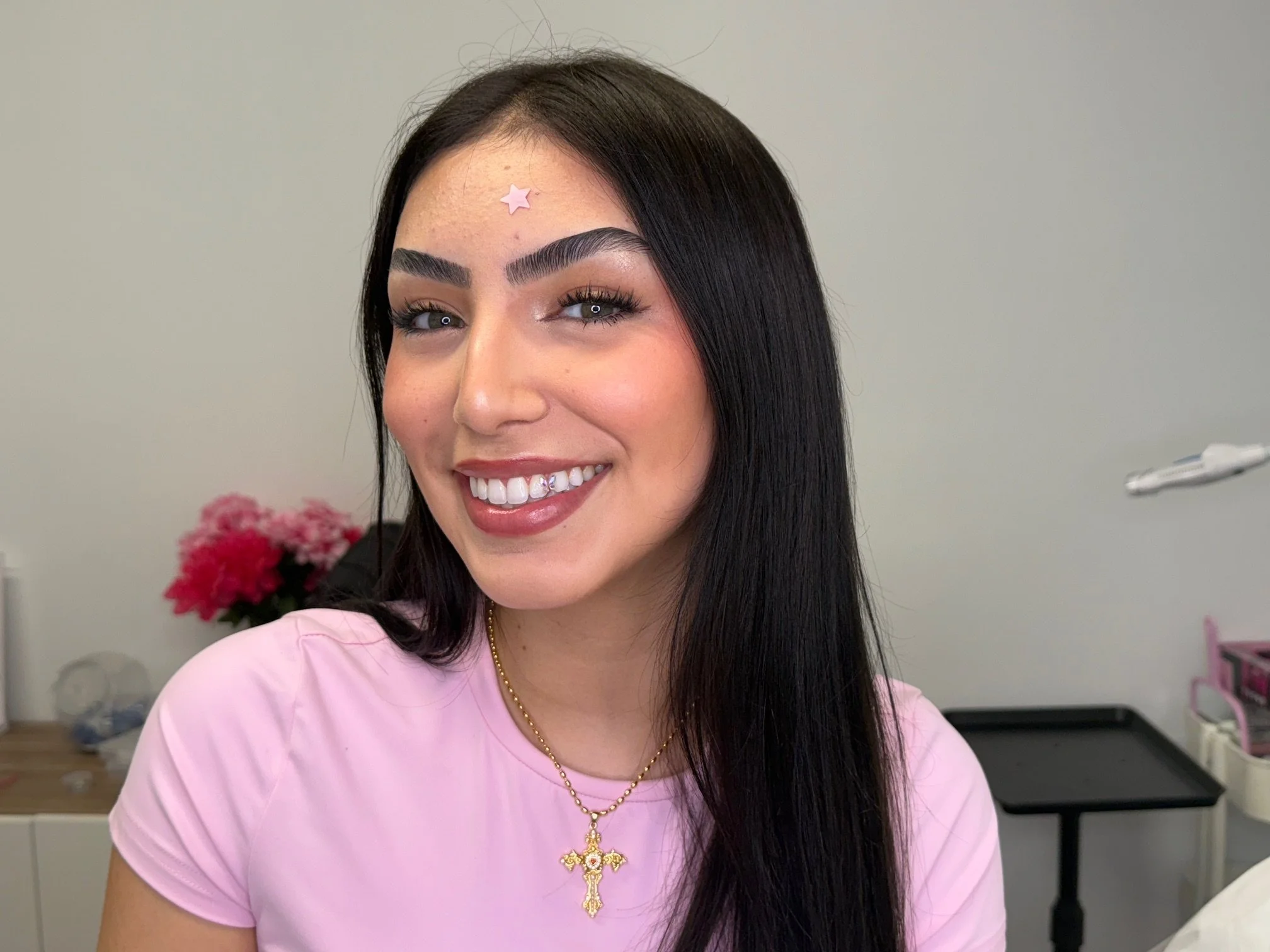A woman with long black hair, light skin, and bright makeup, smiling in a pink shirt with a gold cross necklace. There are flowers and a desk in the background.