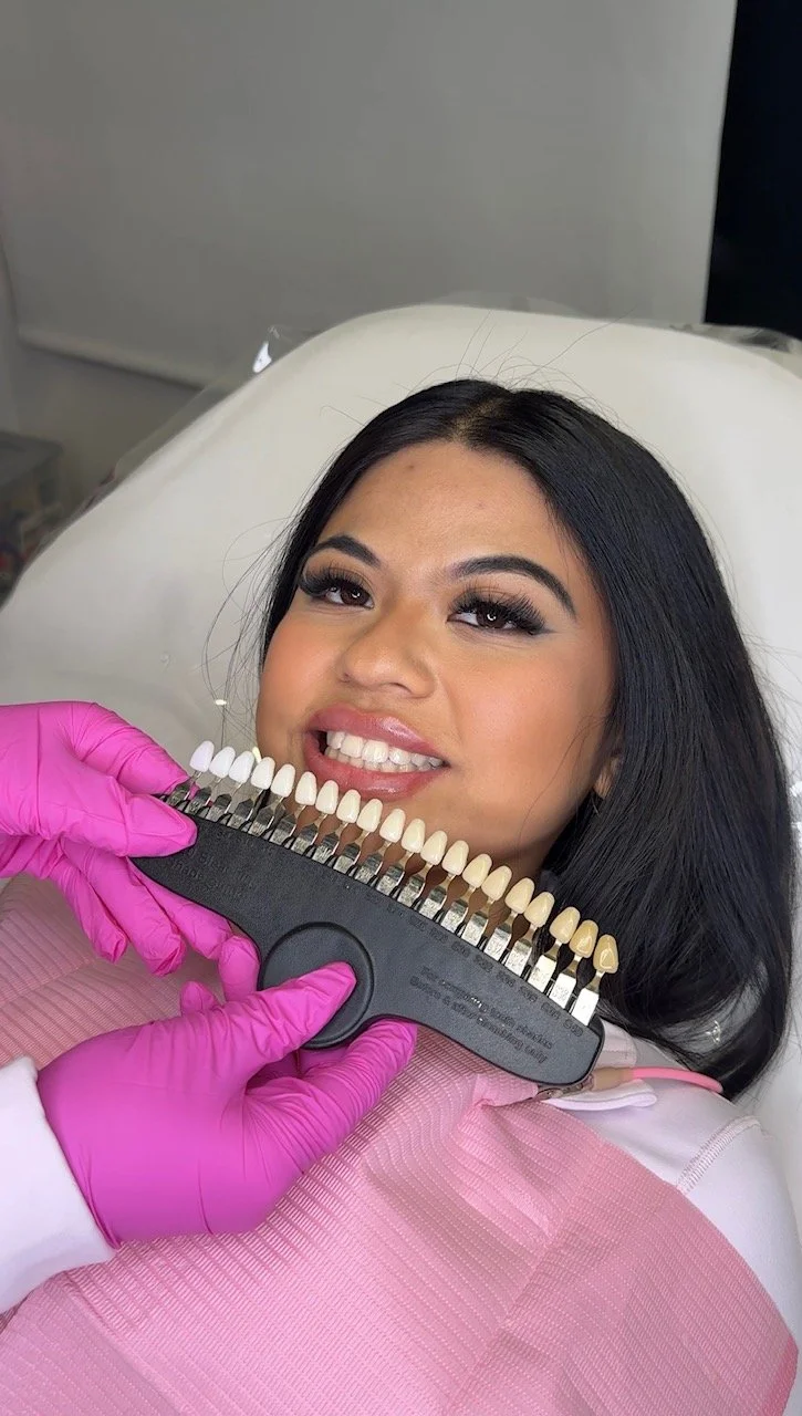 A woman lying on a dental chair smiling while a dental professional shows her a shade guide for teeth color. The woman has dark hair and is wearing makeup, with pink gloves on the dental professional's hand.