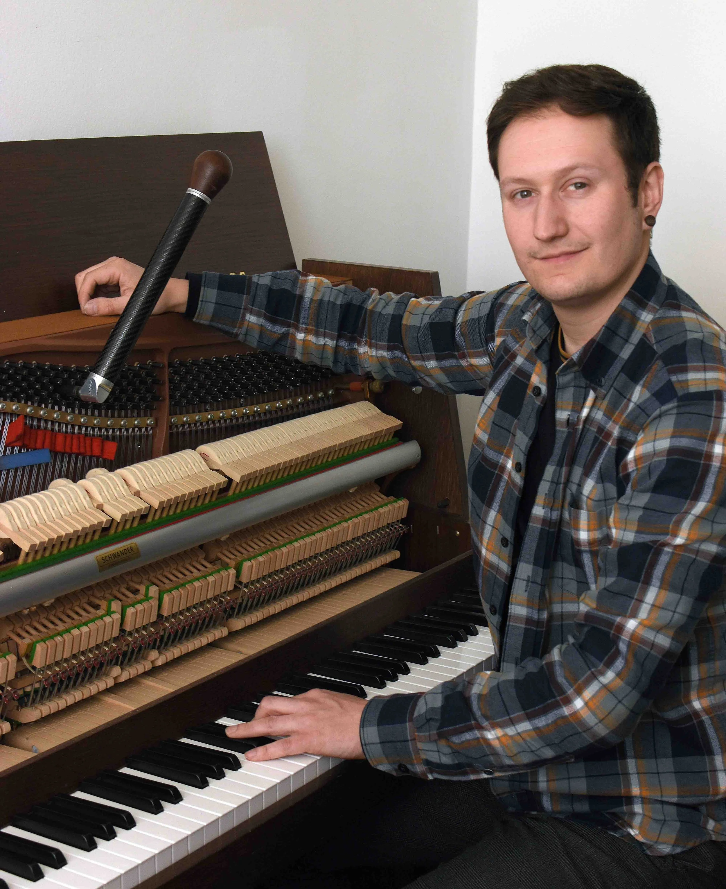 A young man with short dark hair and earring tuning an upright piano, with a hammer resting on the open interior of the piano, wearing a plaid shirt.