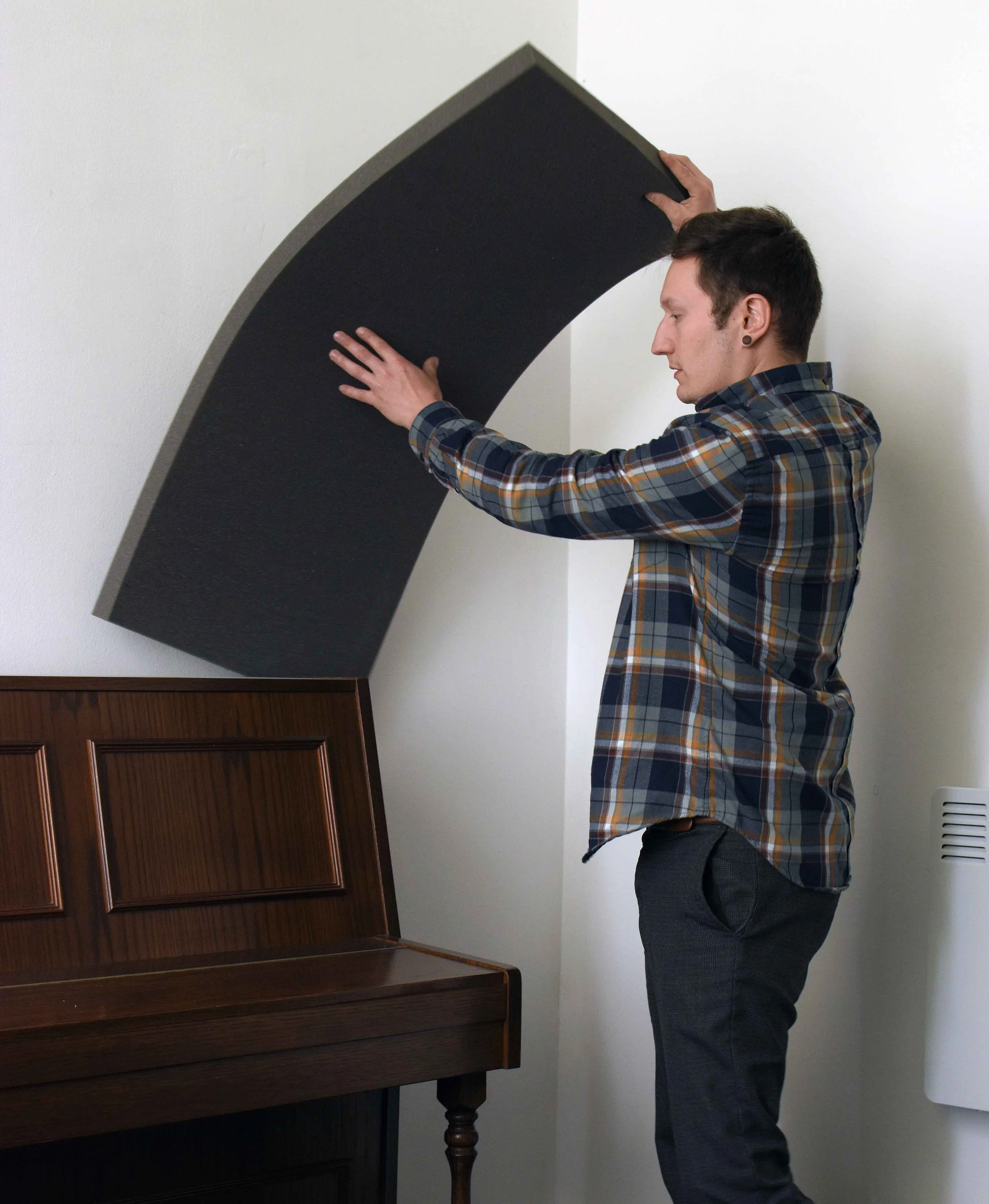 A man in a plaid shirt and dark pants is installing acoustic foam panels as a soundproofing device between an upright piano and the wall behind it.