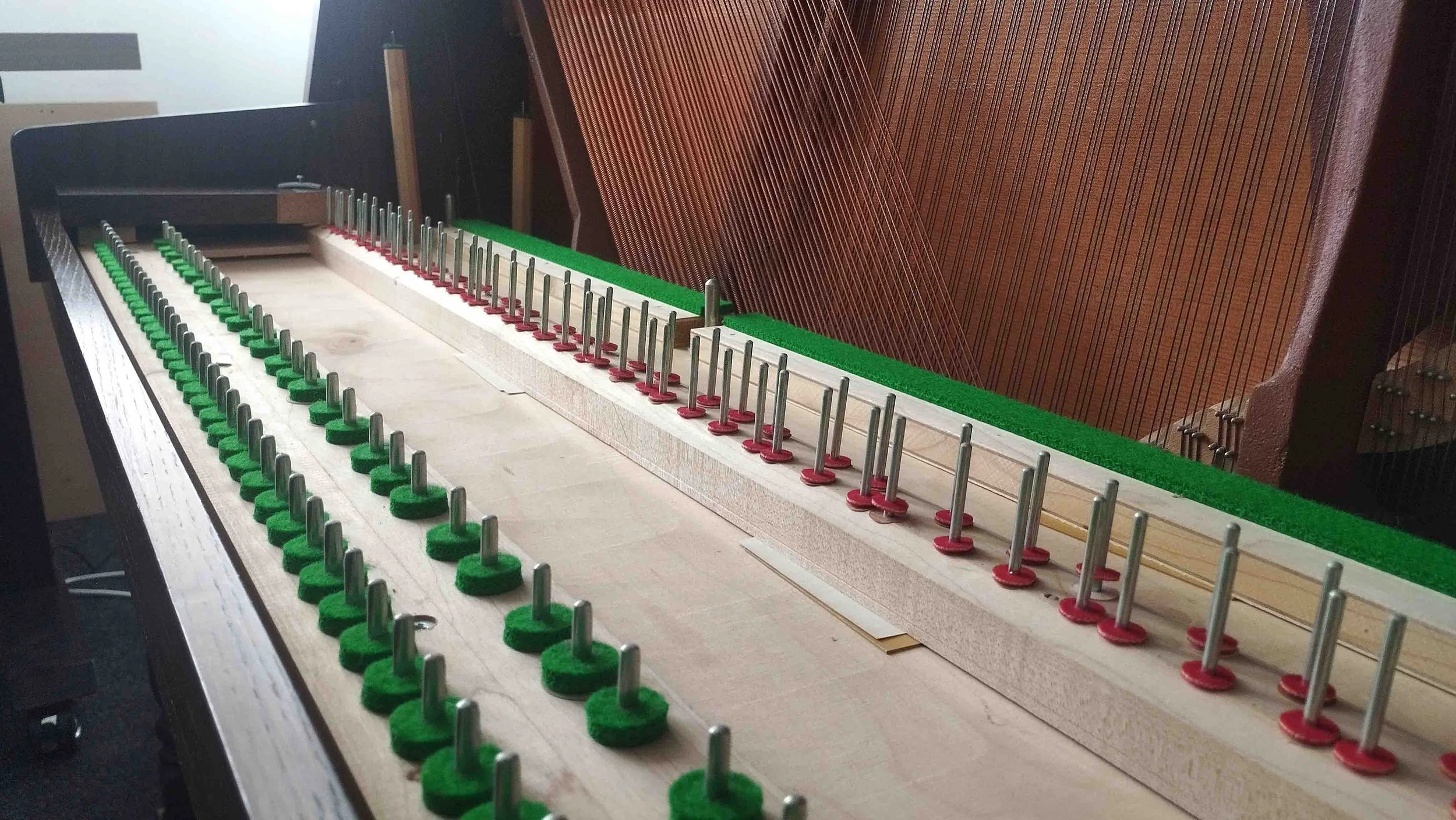 The interior of a piano with its strings and tuning pins exposed, showing the tuning pins in a row with green felt pads around the base.