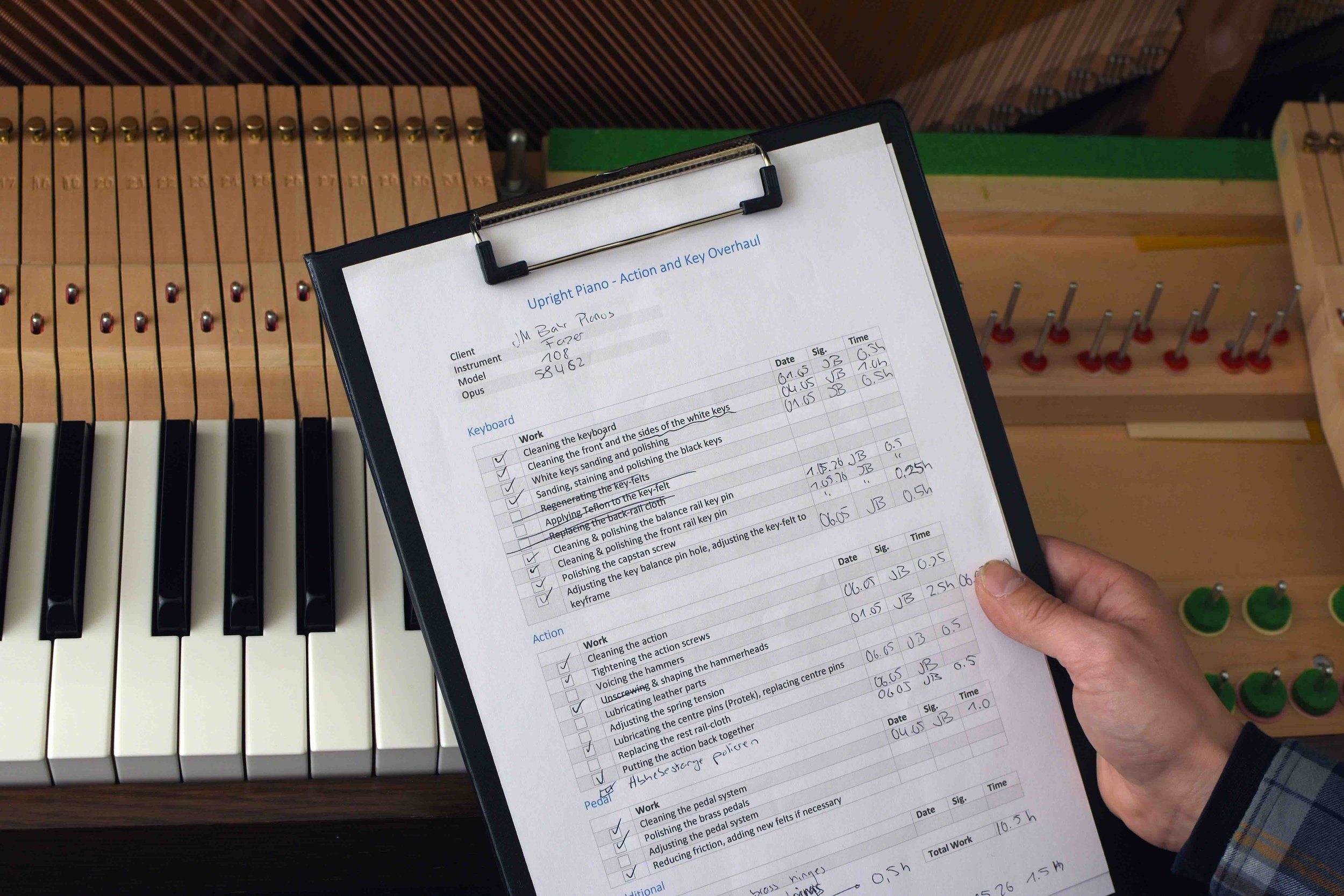 An open sheet of paper with a piano maintenance checklist held by a person over a piano with black and white keys and piano parts, in a workshop environment.