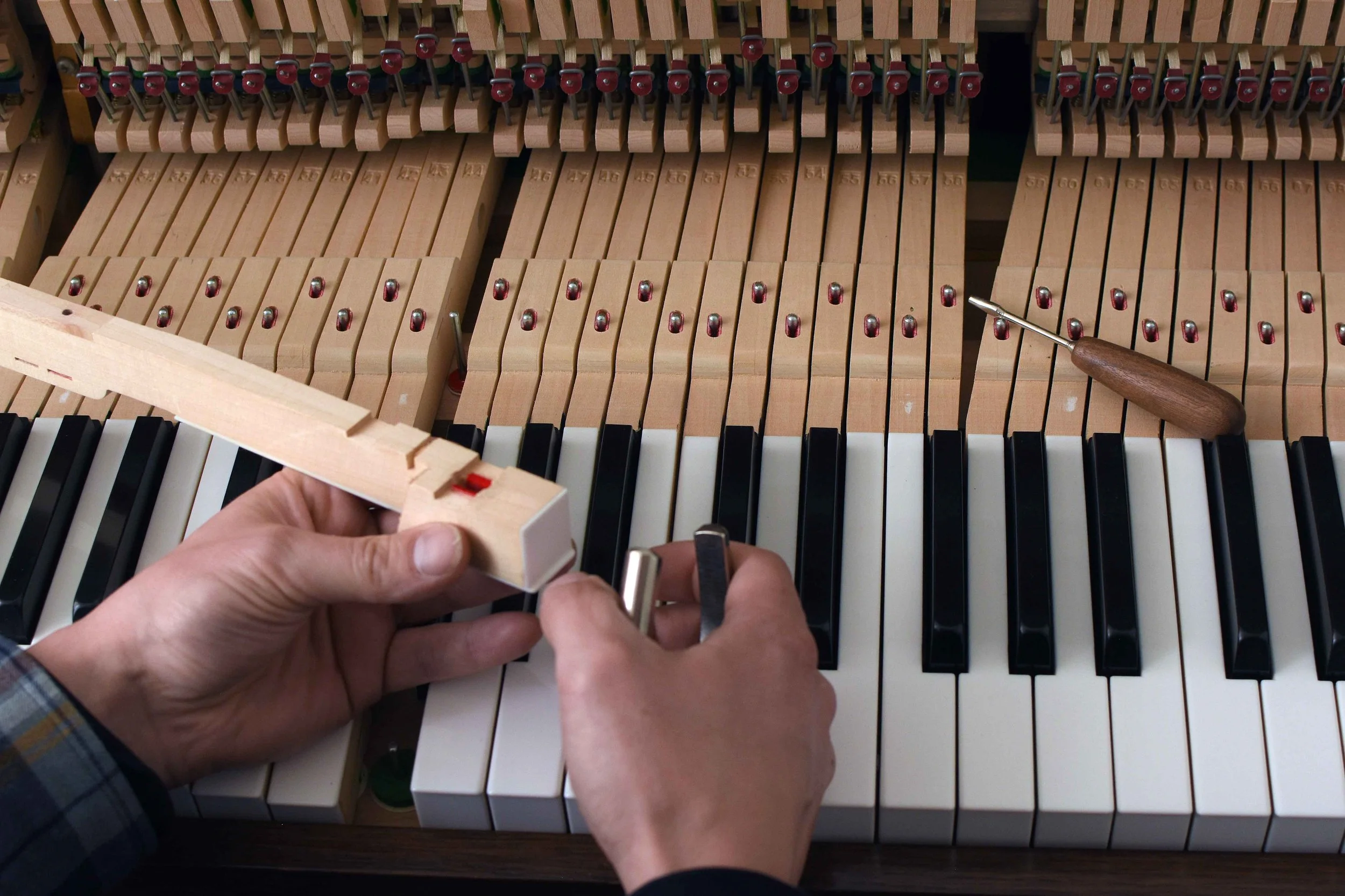 Close-up of person repairing piano with screwdriver and small wooden block inside the piano's open body, showing piano strings and hammers.