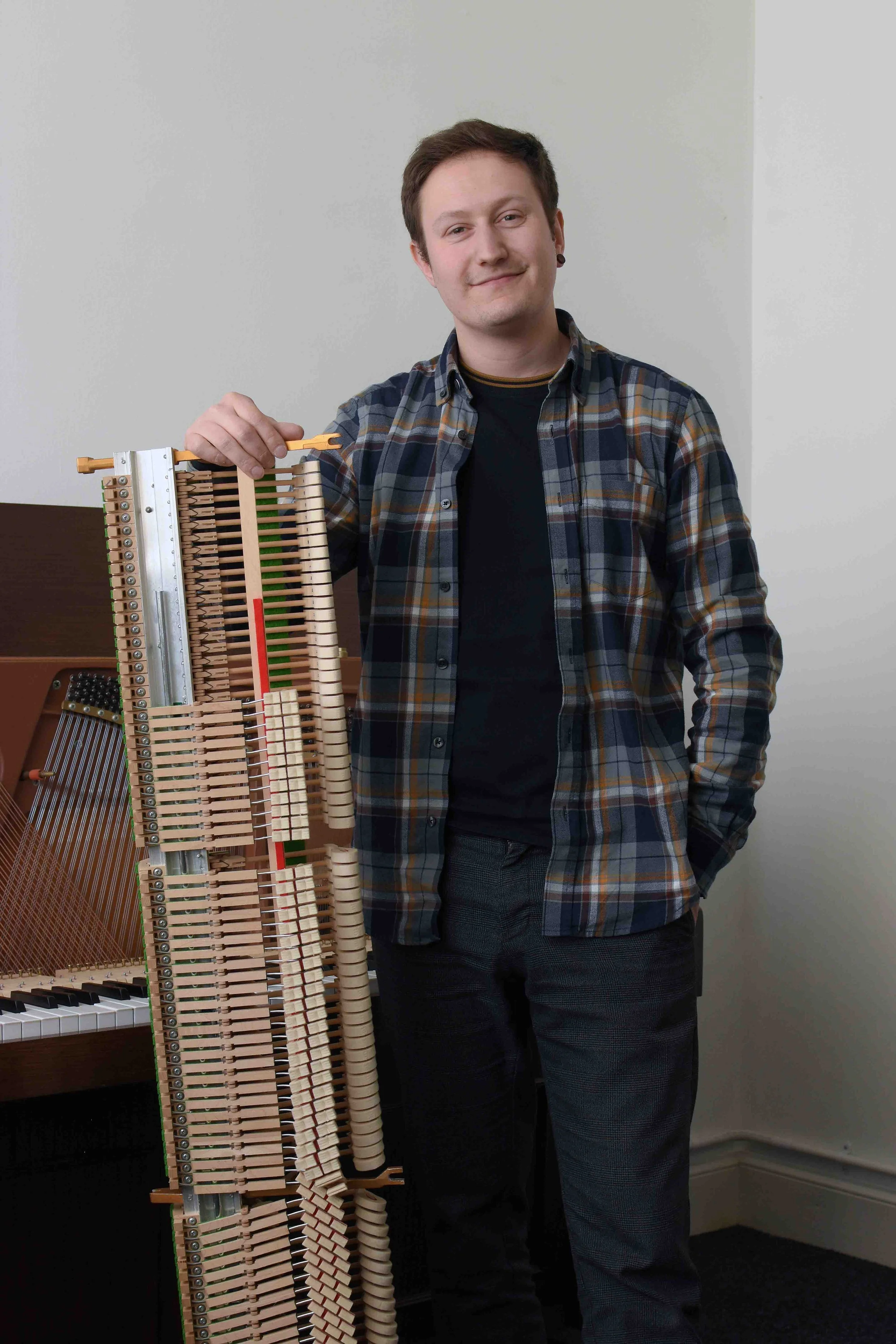 A young man with short brown hair, wearing a plaid shirt and dark trouse, stands next to a partially disassembled upright piano with its internal hammers and strings visible. He is smiling and holding a small tool, in a room with light-colored walls.