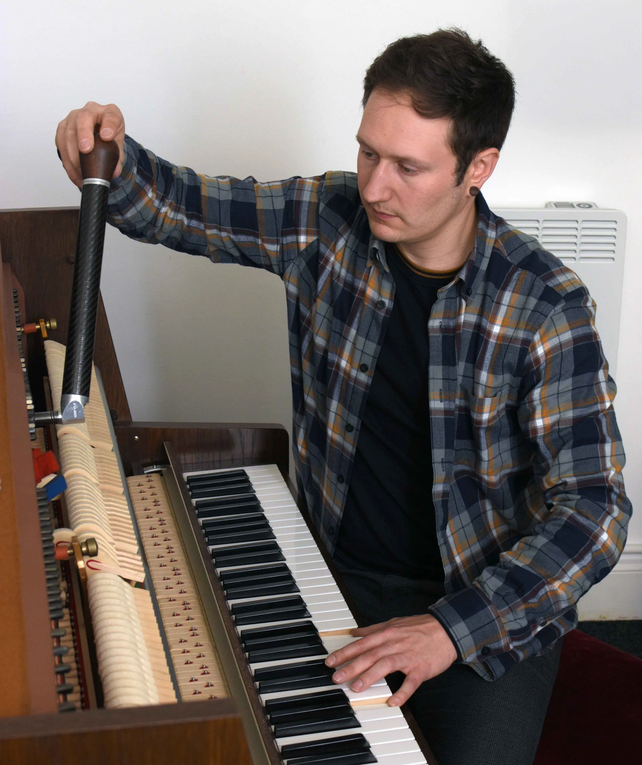 A man in a plaid shirt tunes an upright piano, focusing on his hands and face while pressing keys.
