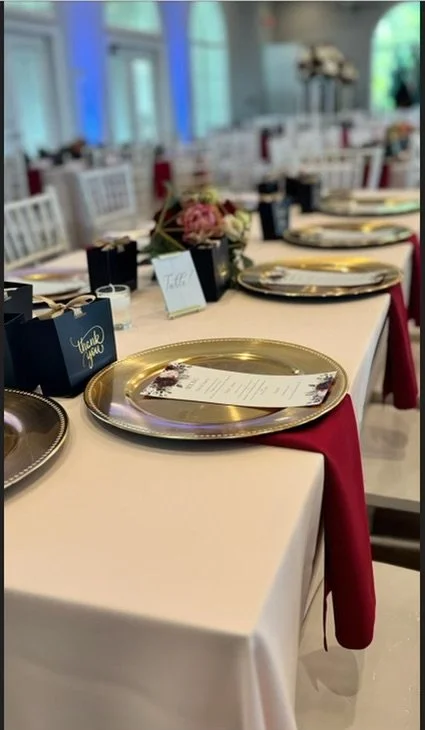Elegant banquet table set for a celebration with gold chargers, navy blue gift boxes, floral centerpieces, and place cards in a bright, spacious room.